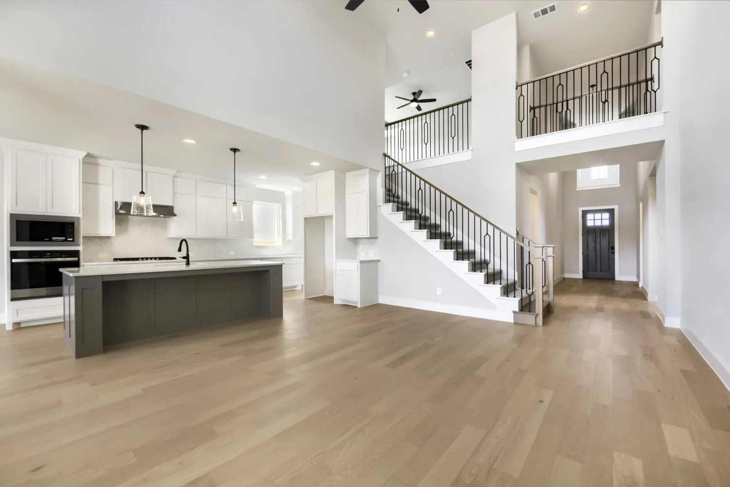 Expansive open-concept living space with wood-finish flooring, a two-story ceiling, and a modern kitchen featuring a large island, stainless steel appliances, and pendant lighting