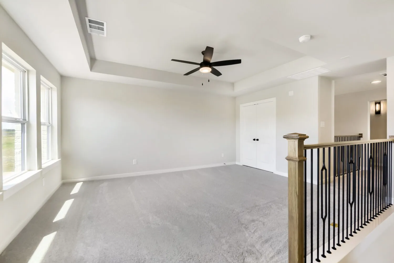 Tray ceiling, ceiling fan, multiple windows, carpet flooring, and a railing with wood newel post and wrought iron spindles