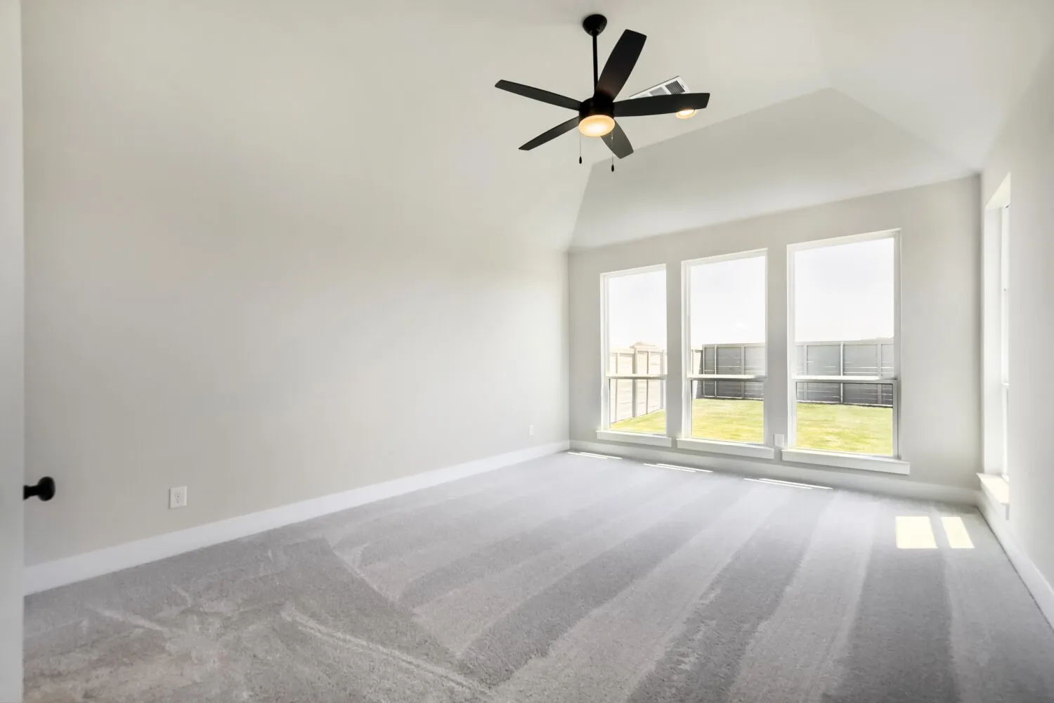 Vaulted ceiling with a black ceiling fan, multiple windows with backyard views, and light gray carpeting