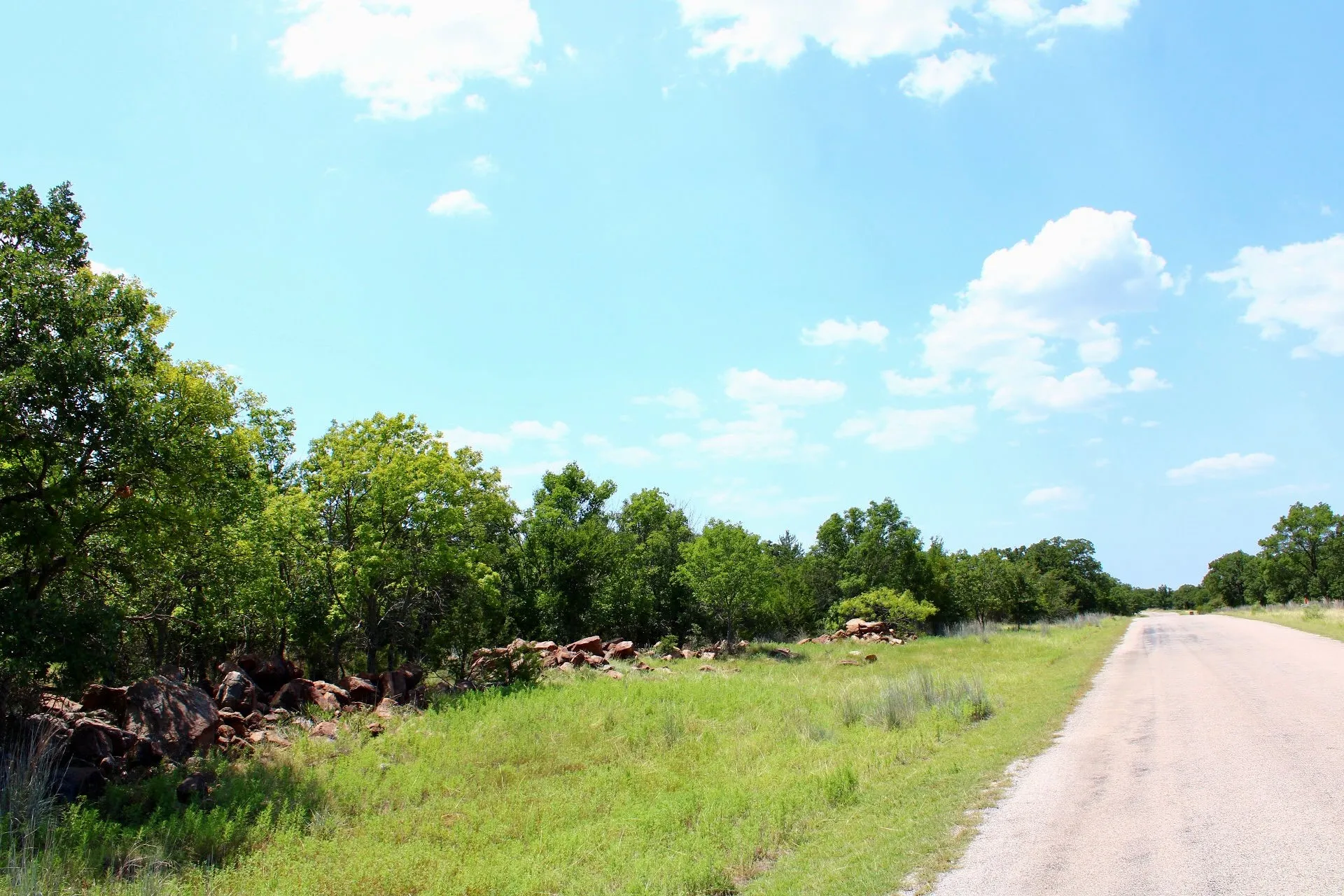 Paved road bordering a tree-lined property with natural rock features and green grass