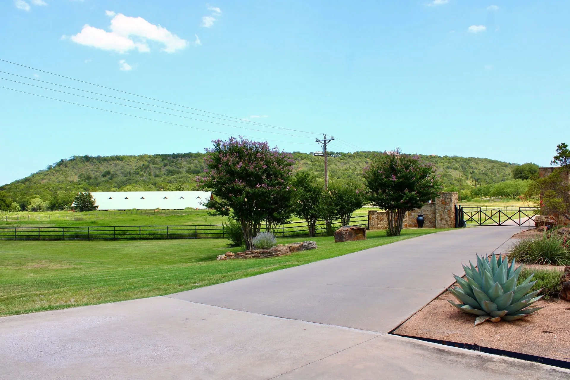 Paved driveway leading to a stone entry gate, featuring mature landscaping with agave plants and blooming crepe myrtles