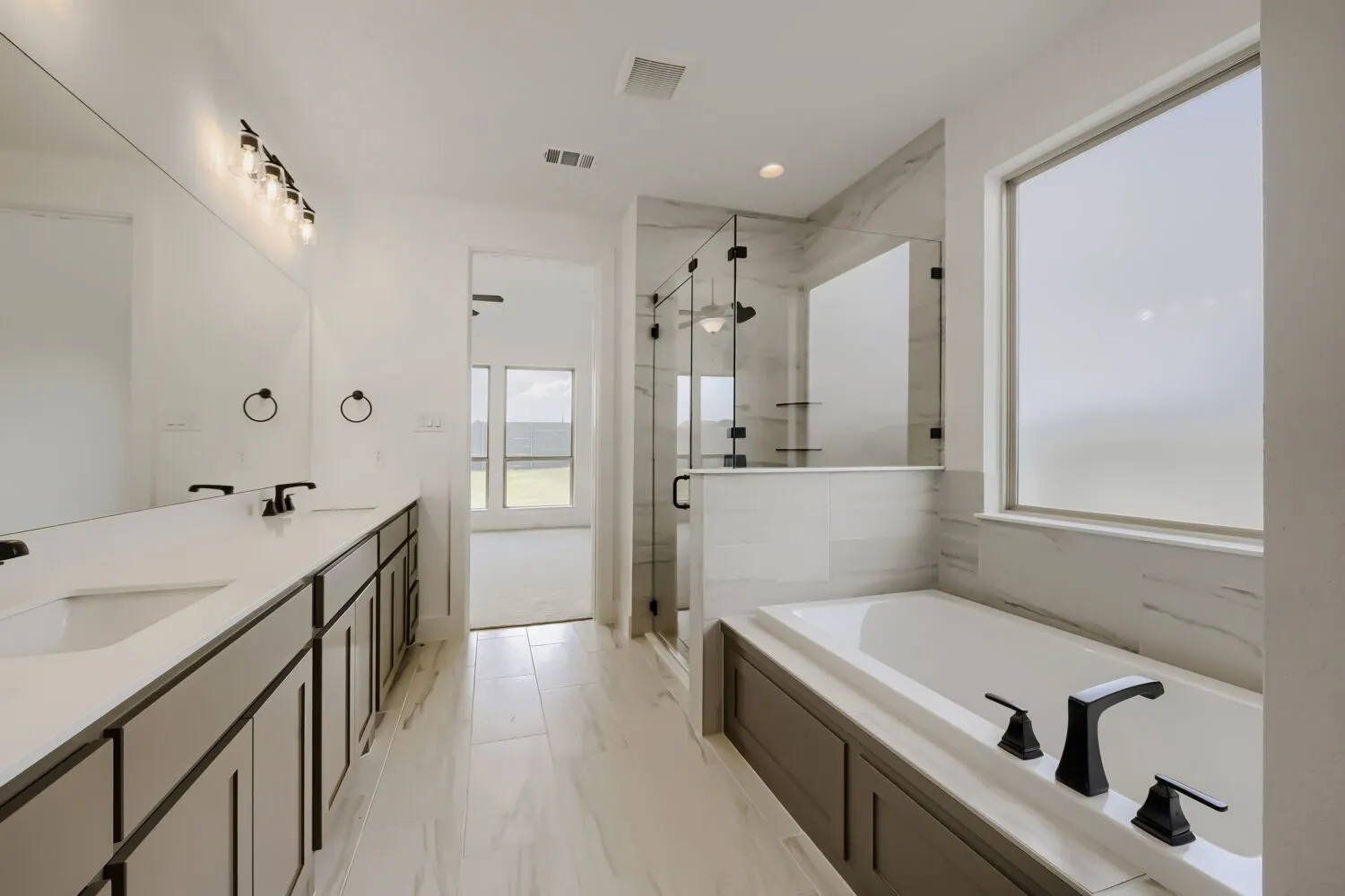 Spa-inspired primary bathroom featuring dual vanities with wood-finish cabinetry and quartz countertops, a freestanding soaking tub with matte black fixtures, a walk-in shower with frameless glass enclosure and subway tile surround, heated floor, and abundant natural light from oversized windows