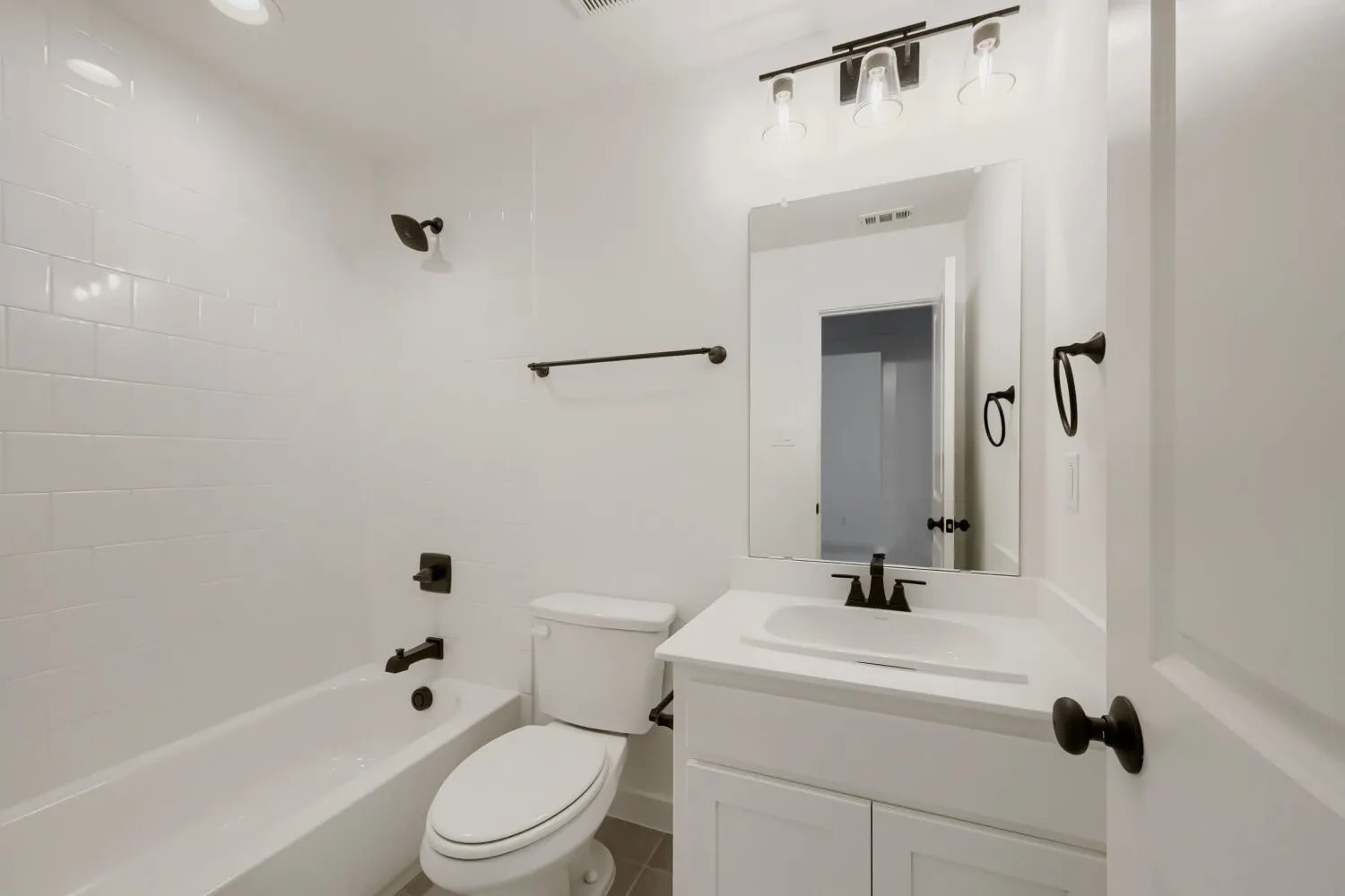 White subway tile shower surround, matte black fixtures, white vanity with integrated sink, rectangular vanity mirror, and ceiling-mounted lighting