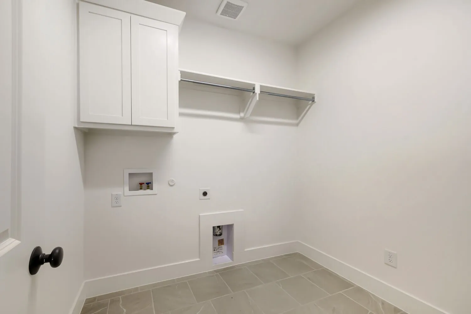 White shaker-style cabinetry, overhead shelving, and floor-level utility hookups, all complemented by light-toned floor tile