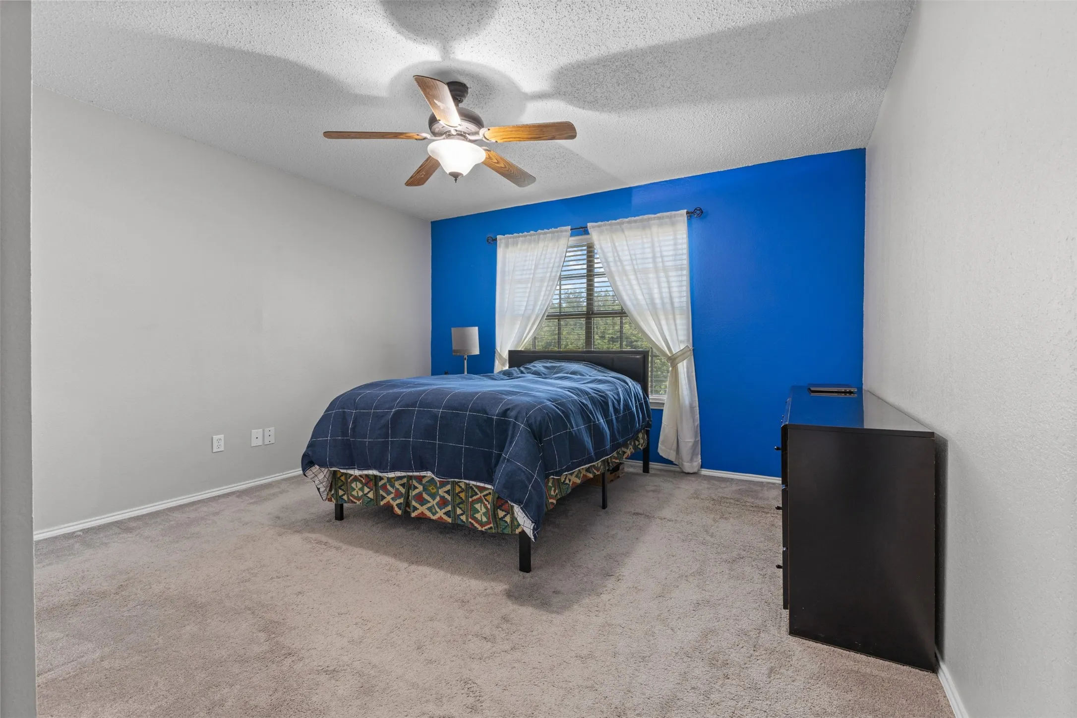 Carpeted interior space featuring a ceiling fan with integrated lighting, a window with blinds and sheer curtains, and a prominent accent wall