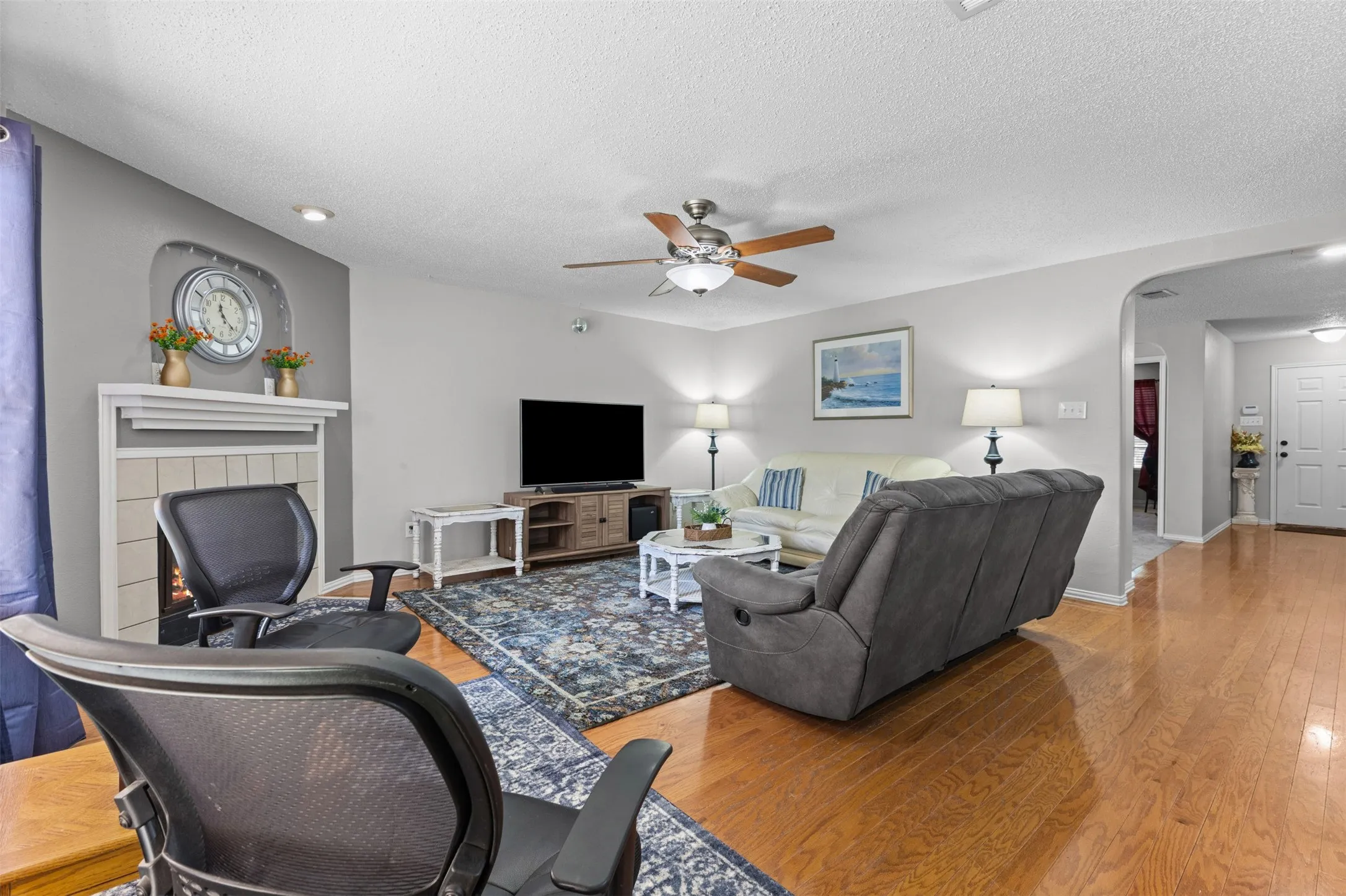 Living area featuring wood-finish flooring, a ceiling fan with integrated lighting, and a fireplace with a white mantel and tiled surround