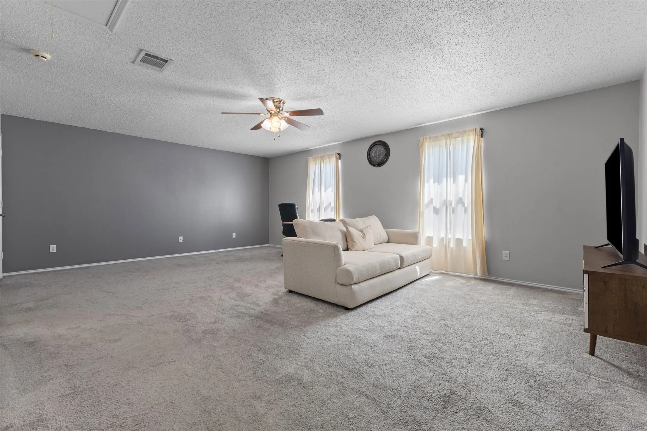 Spacious room featuring light gray carpeting, a two-tone wall aesthetic with a dark accent wall, a ceiling fan with integrated lighting, and multiple windows providing ambient illumination