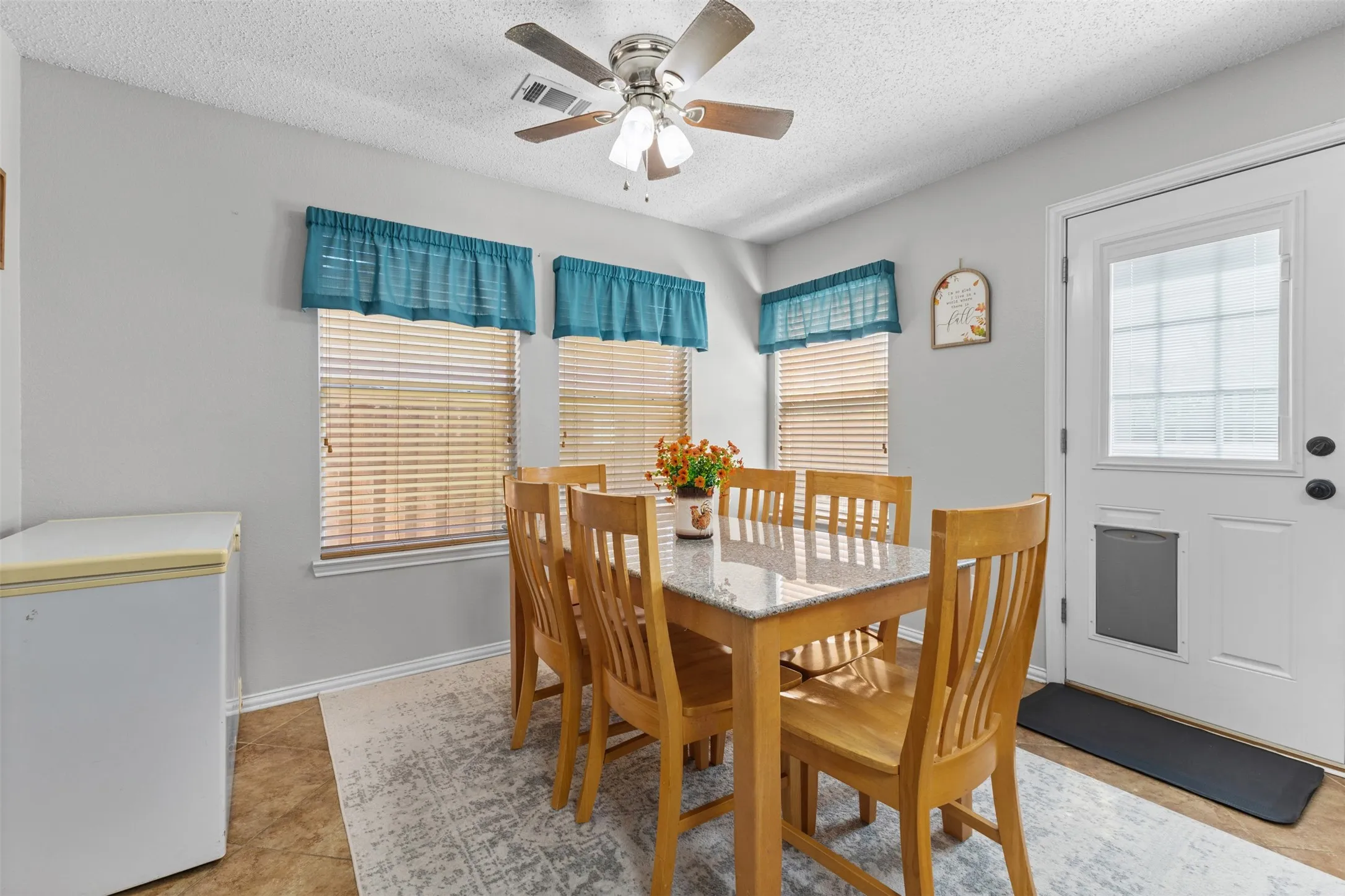 Dining area featuring a ceiling fan with light fixture, three windows with blinds, a white exterior door with glass insert, and tile flooring