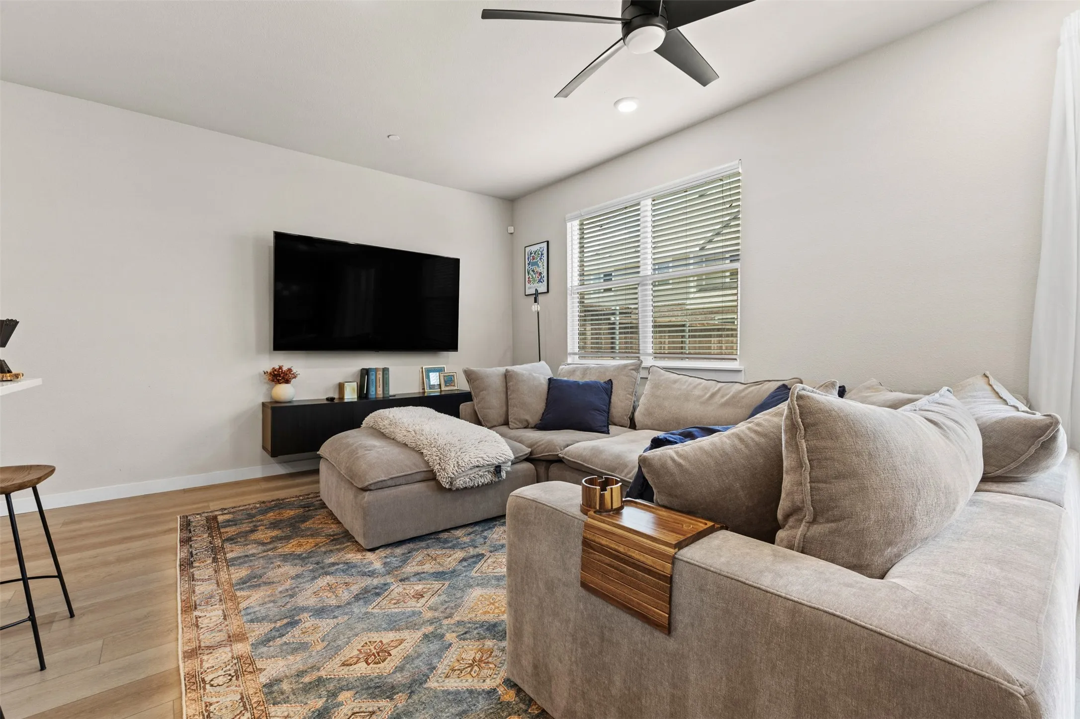 Living area featuring light wood-finish flooring, a wall-mounted television, a floating media console, a ceiling fan with integrated lighting, and a large window with blinds