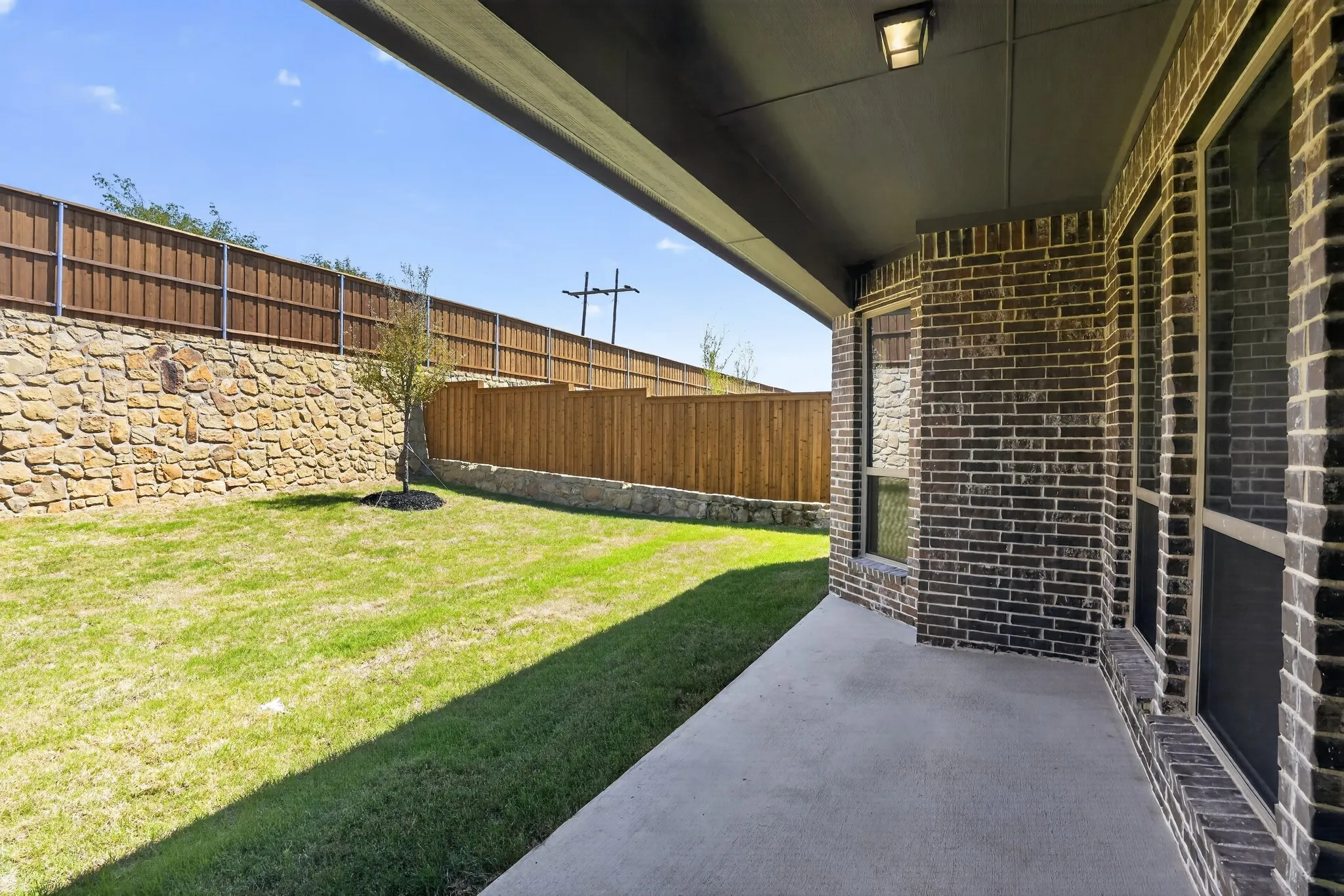 Covered patio with concrete flooring, dark brick exterior, multiple windows, and a landscaped backyard featuring a stone retaining wall and wooden fencing