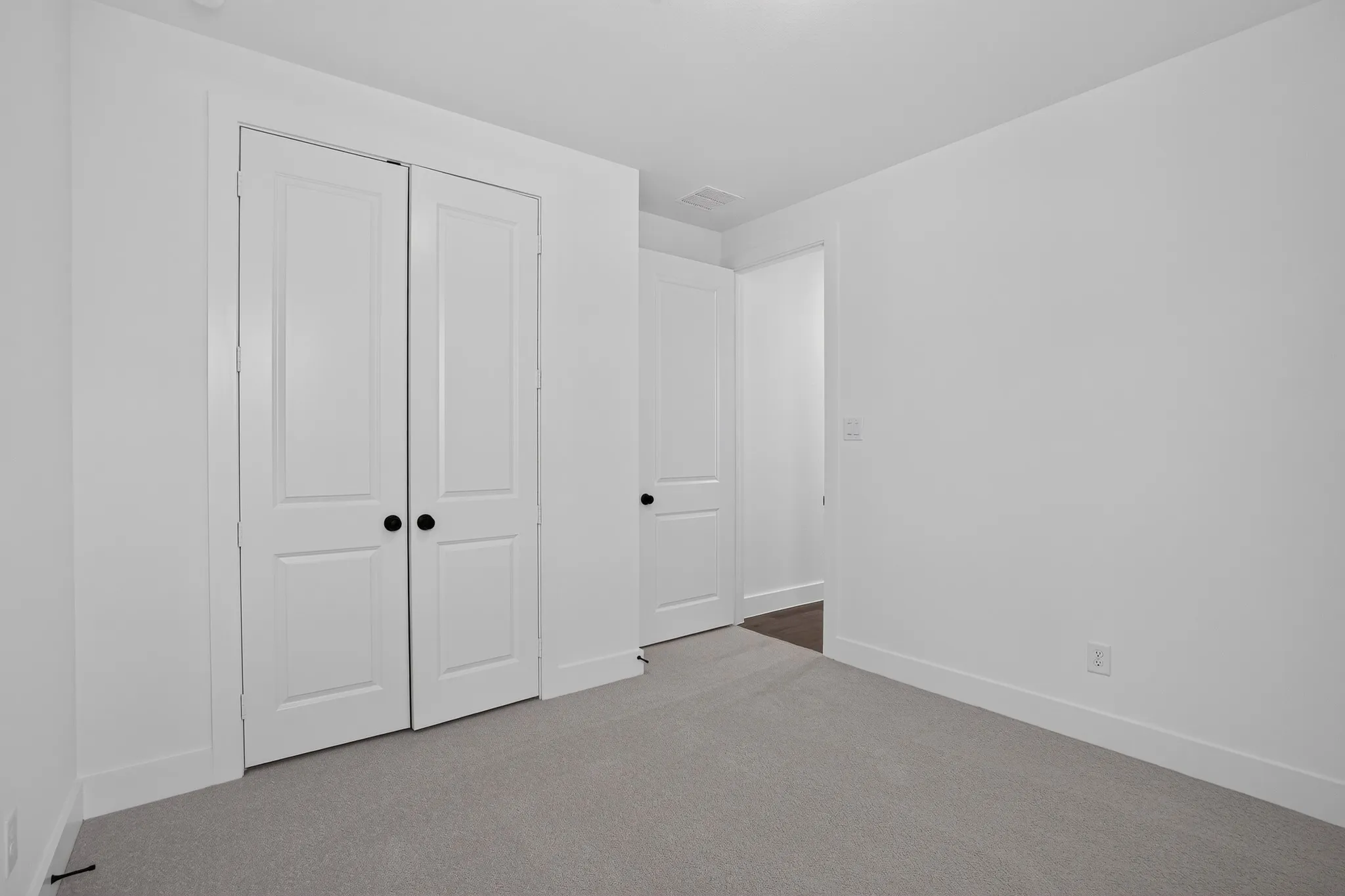 Light gray carpeted flooring, white painted walls, white bifold closet doors with black hardware, and white trim