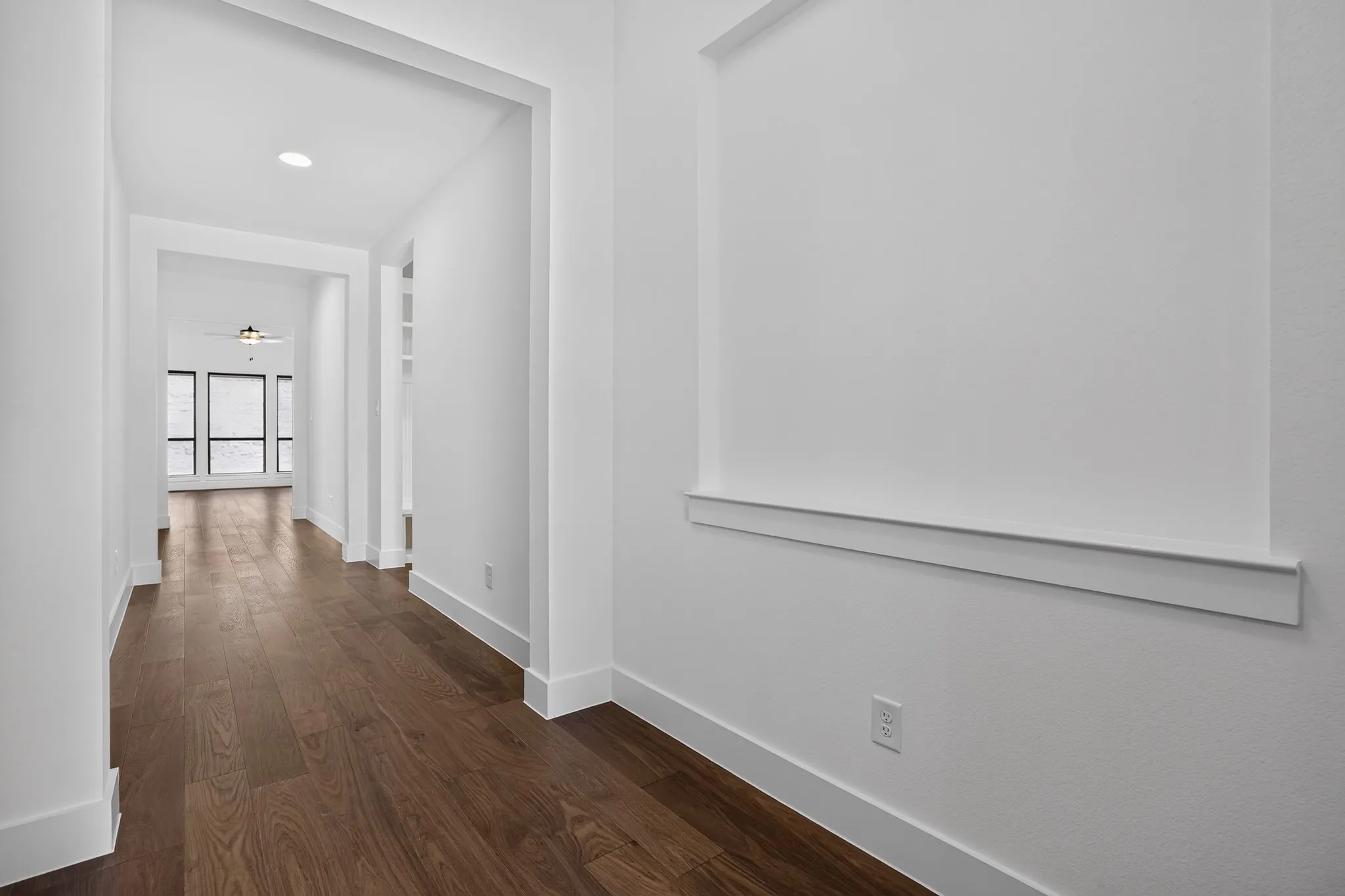Bright hallway featuring rich wood-finish flooring, clean white walls with chair rail molding, recessed lighting, and multiple doorway openings that define the circulation space