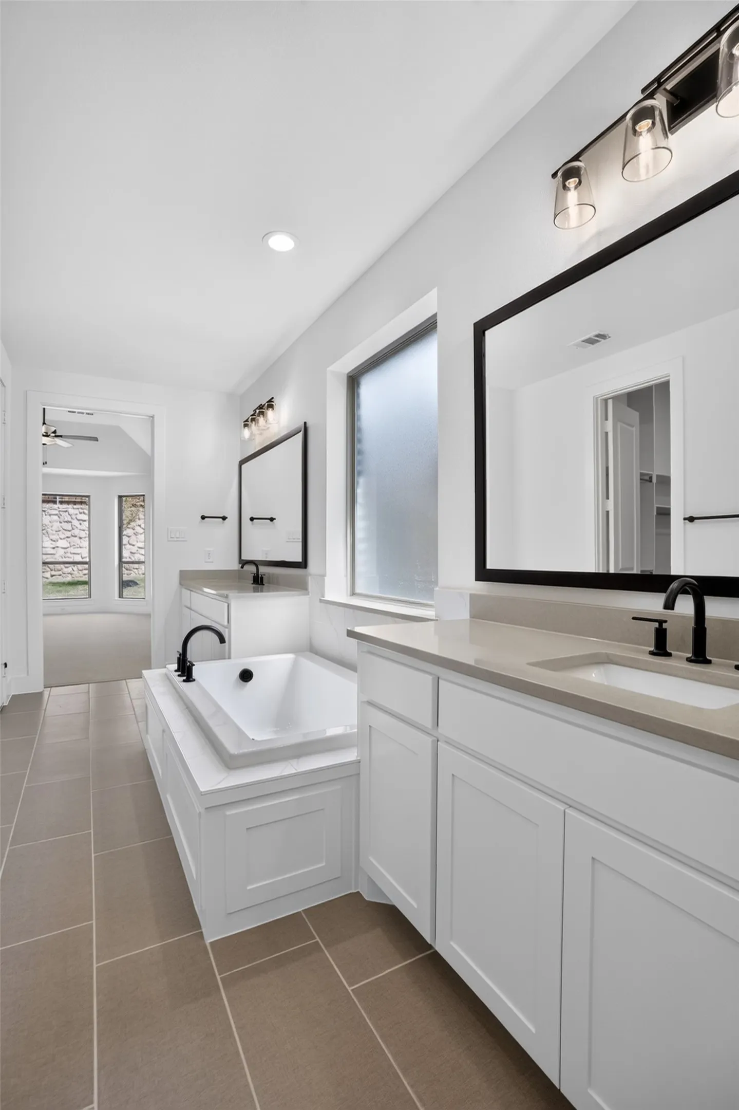 Built-in soaking tub, dual vanities with light countertops, black-framed mirrors, and rectangular tile flooring