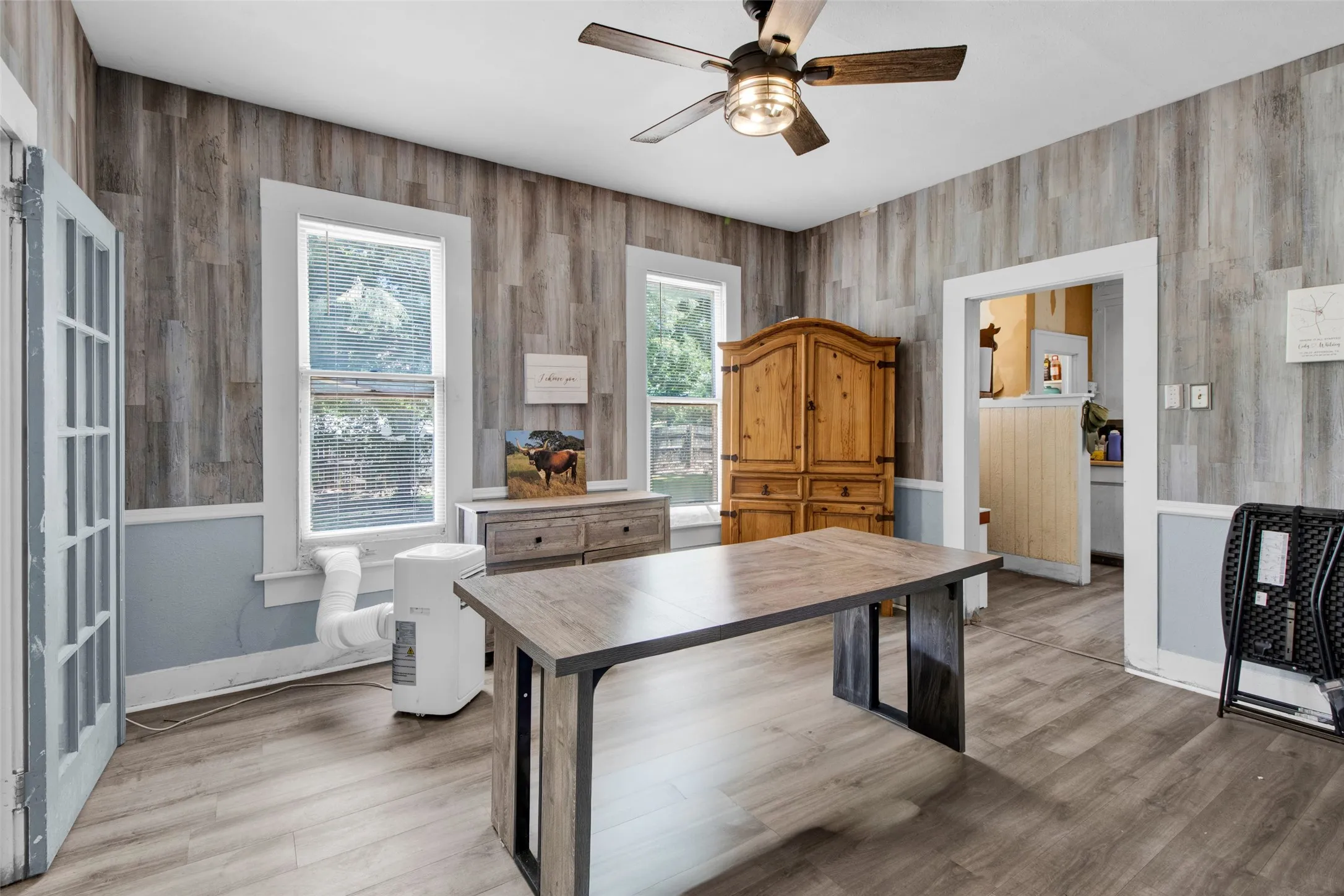 Room featuring wood-finish flooring, plank wall paneling, painted lower wall trim, a ceiling fan with integrated lighting, and multiple windows