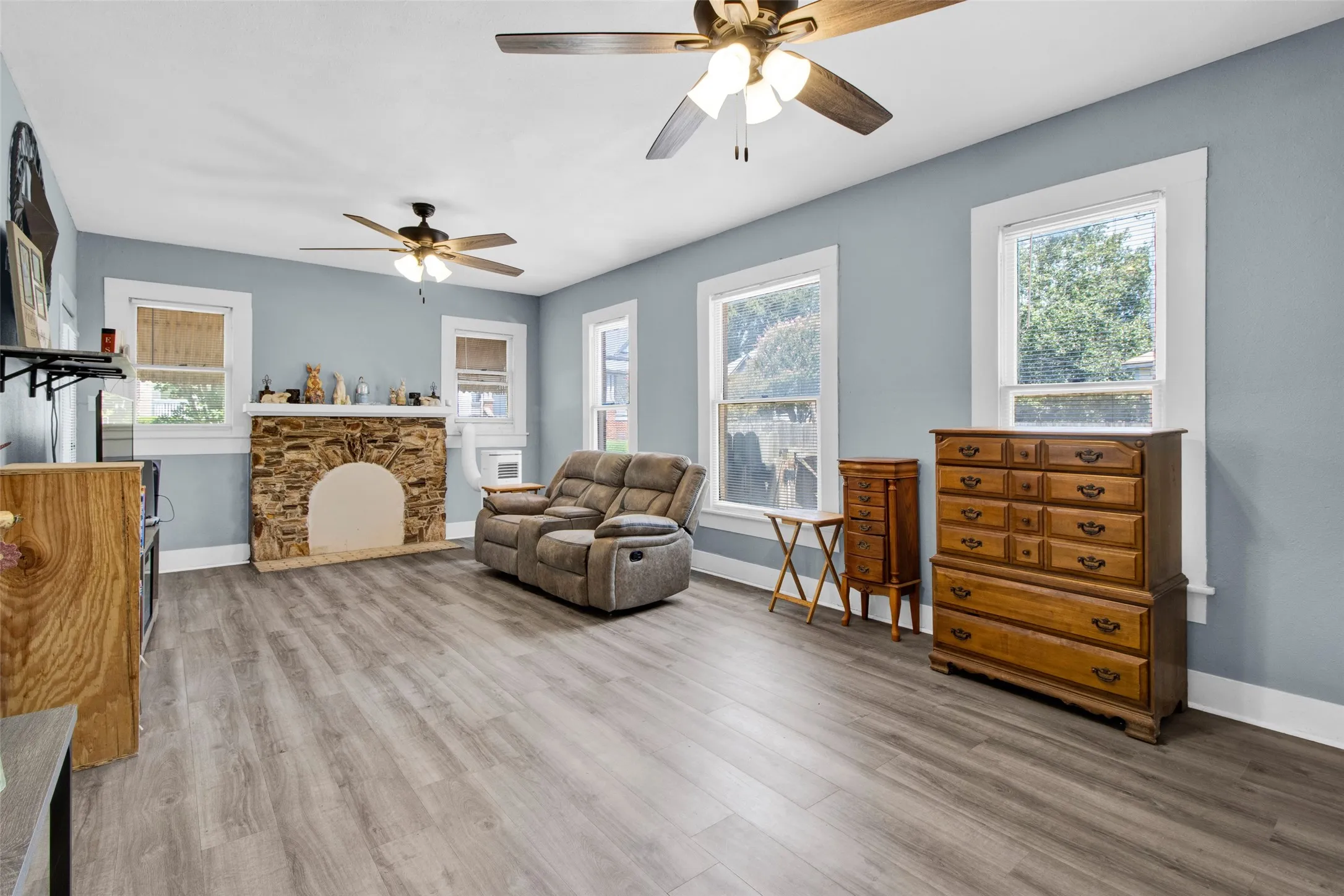 Spacious room featuring wood-finish flooring, a stone-clad fireplace with an arched opening, multiple windows with white trim, and two ceiling fans
