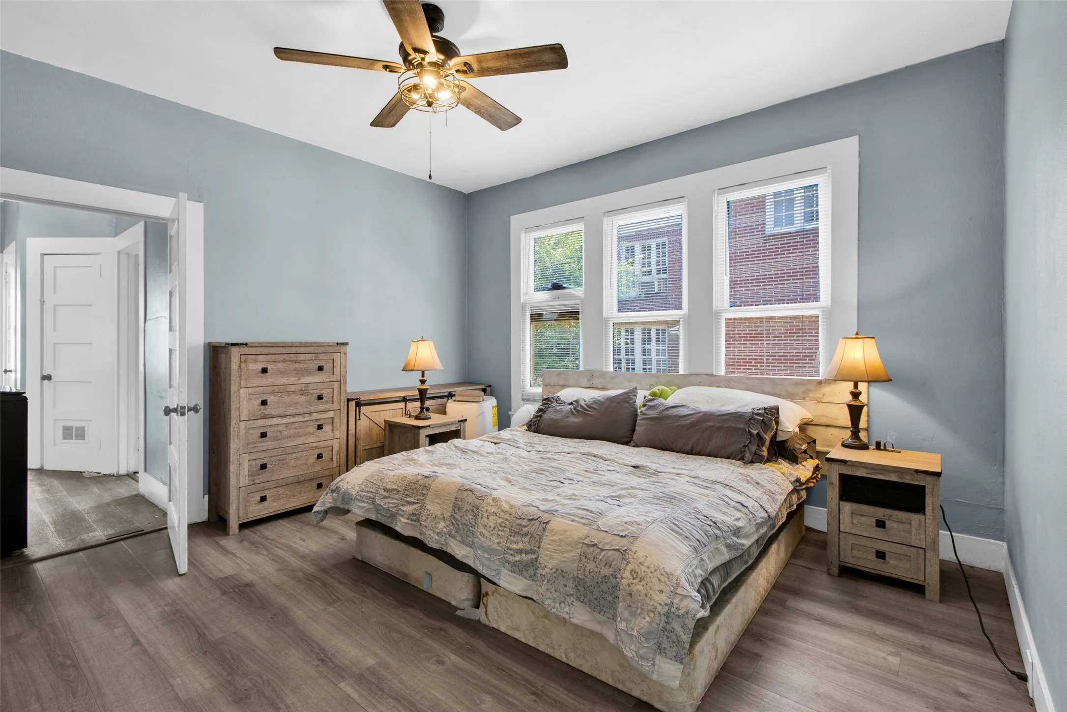 Interior room featuring three sash windows, wood-finish flooring, light blue wall paint, a ceiling fan with integrated lighting, and white trim