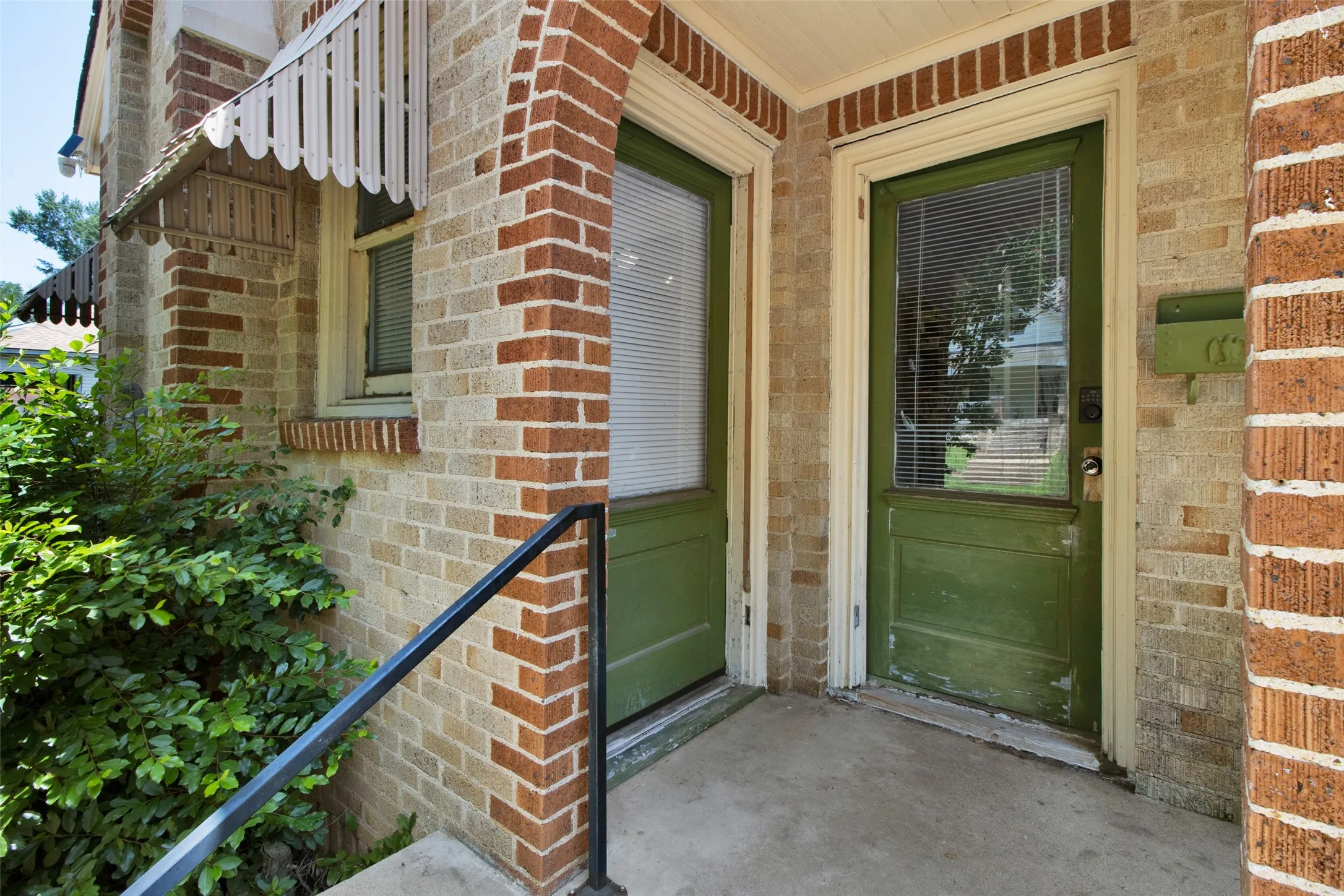 Brick exterior featuring a concrete porch, two green entry doors with glass panels, and a metal handrail
