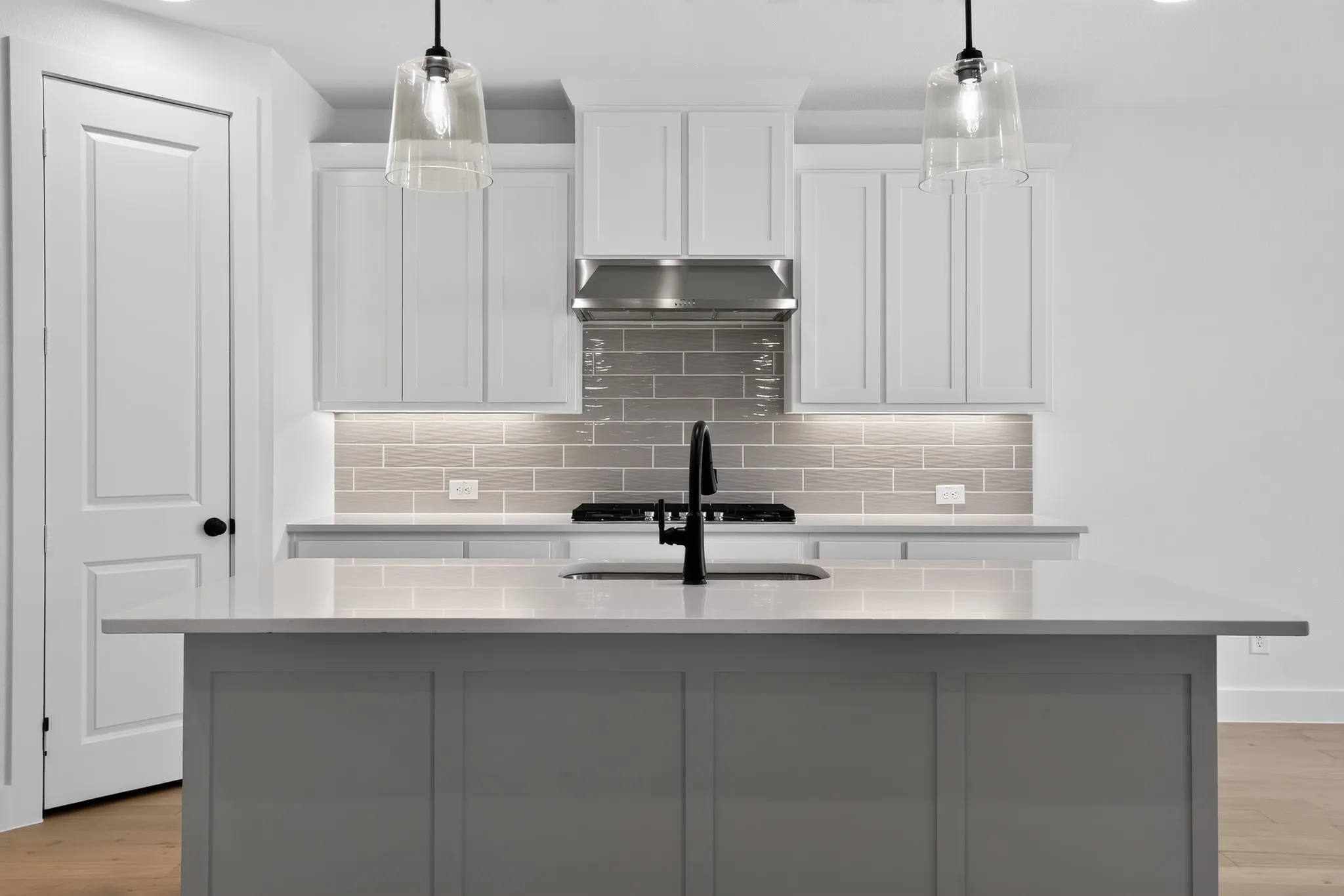 Kitchen featuring an island with a built-in sink, white shaker-style cabinetry, stainless steel range hood, grey subway tile backsplash, and two pendant lights