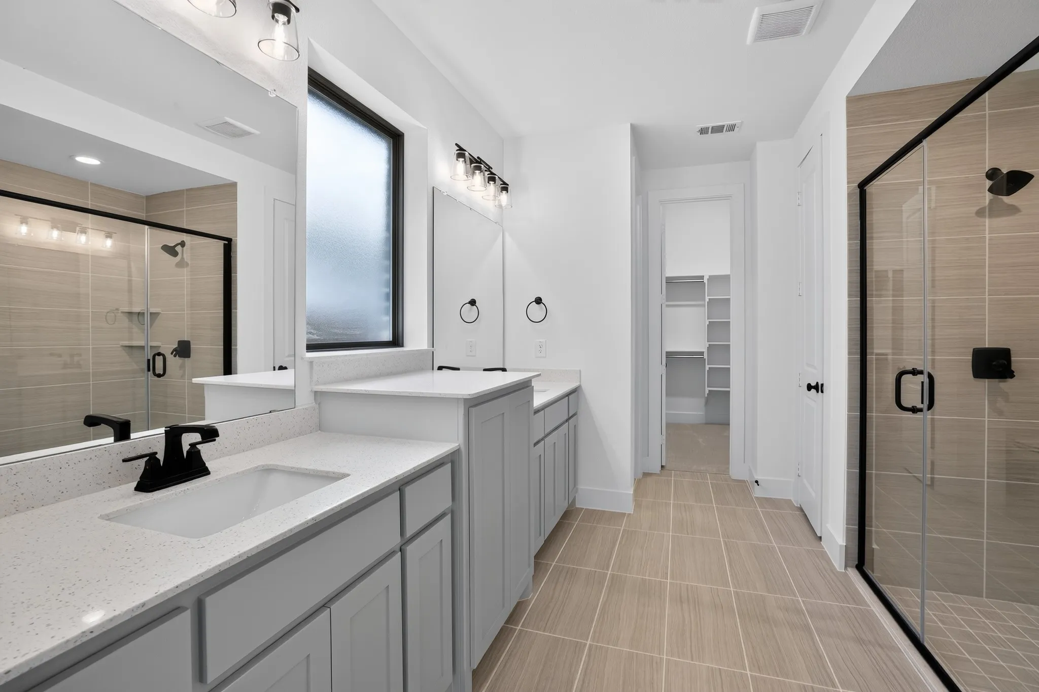 Dual vanities with integrated sinks and white countertops, walk-in shower with glass enclosure and tiled walls, black-finish fixtures, and a walk-in closet with built-in shelving