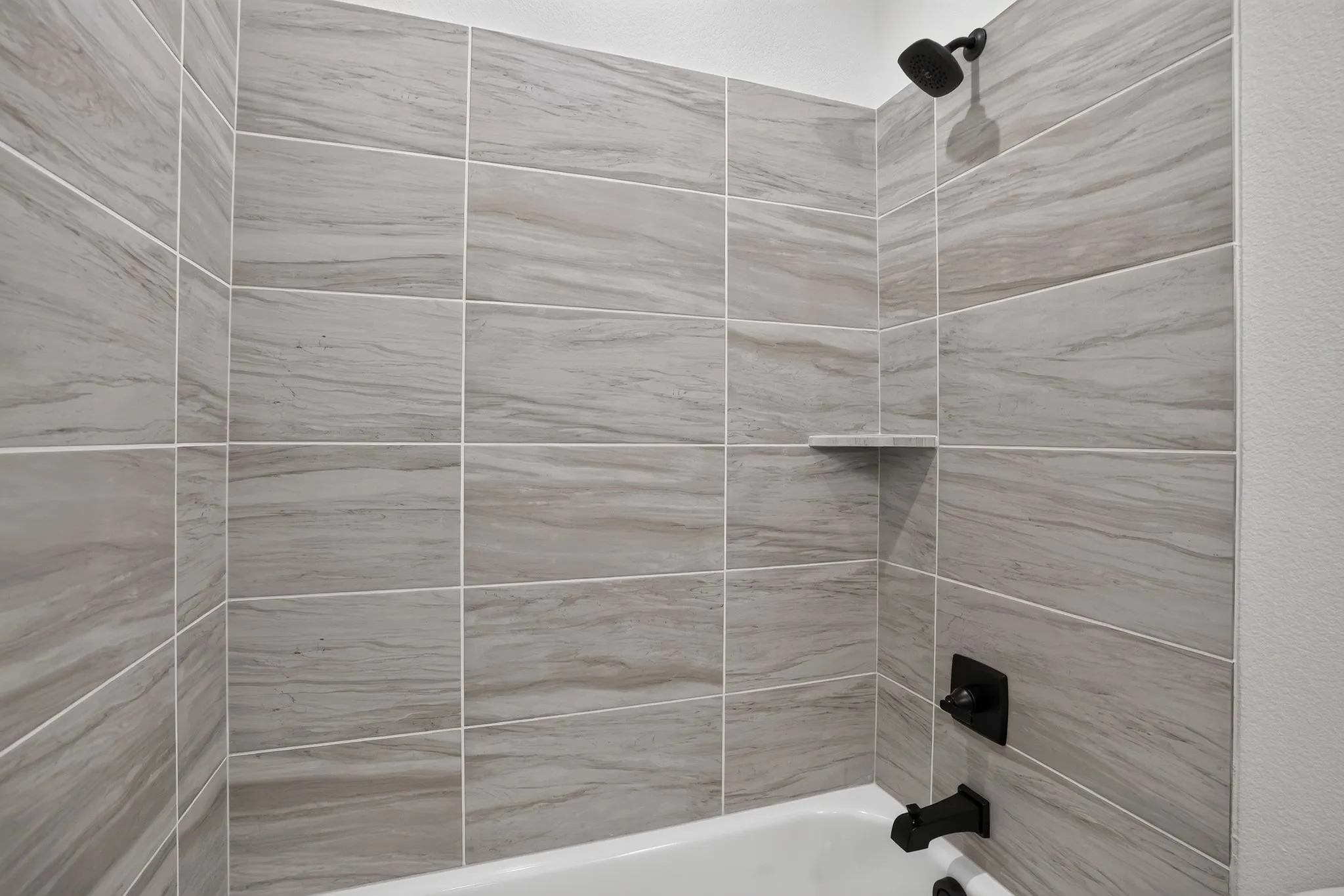 Bathtub and shower combination featuring a tiled surround, integrated shower shelf, and matte black fixtures