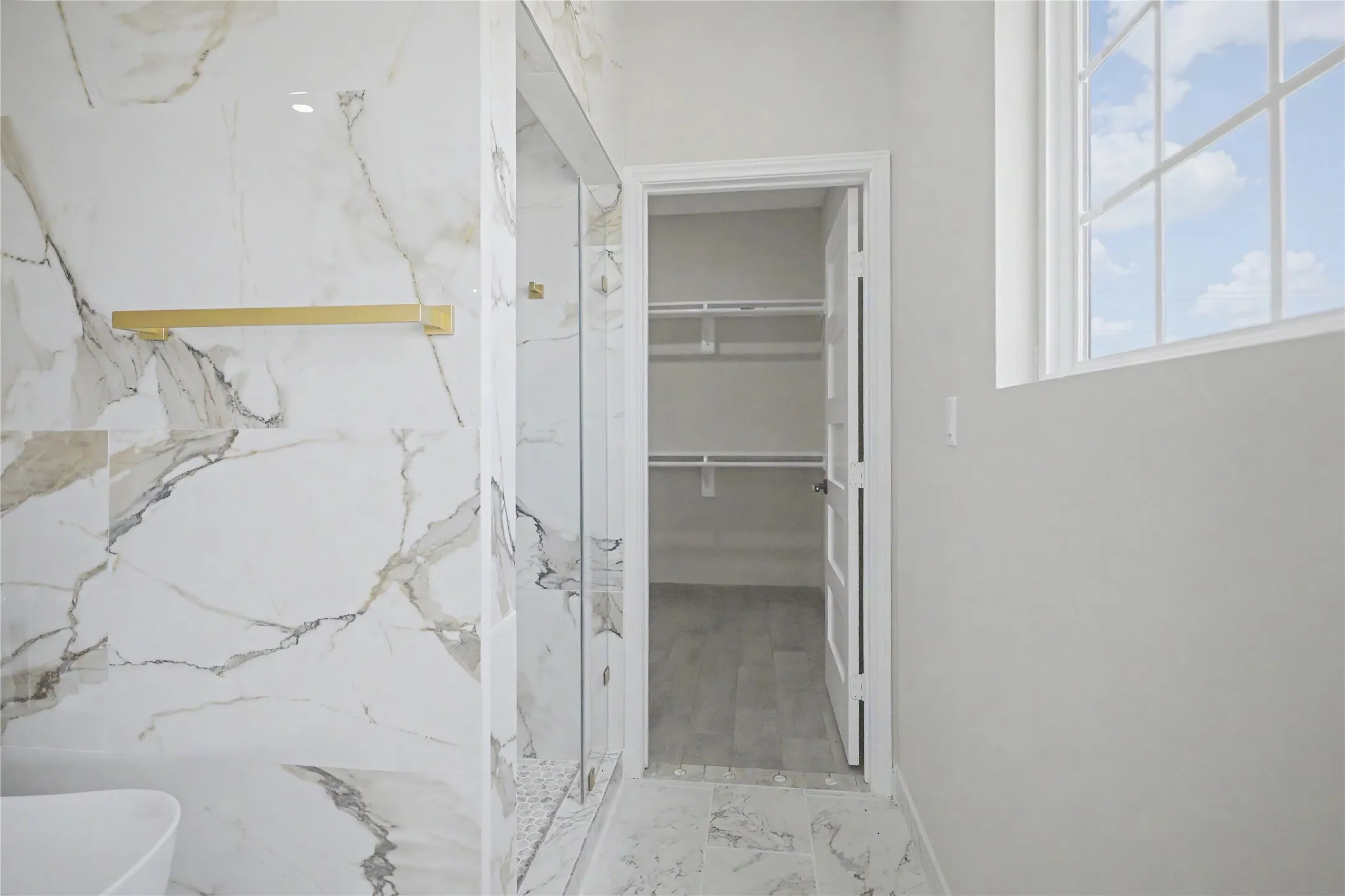 Spacious bathroom featuring large format marble-look tile, a glass-enclosed shower, gold-tone fixtures, a built-in storage closet with shelving, and a multi-pane window