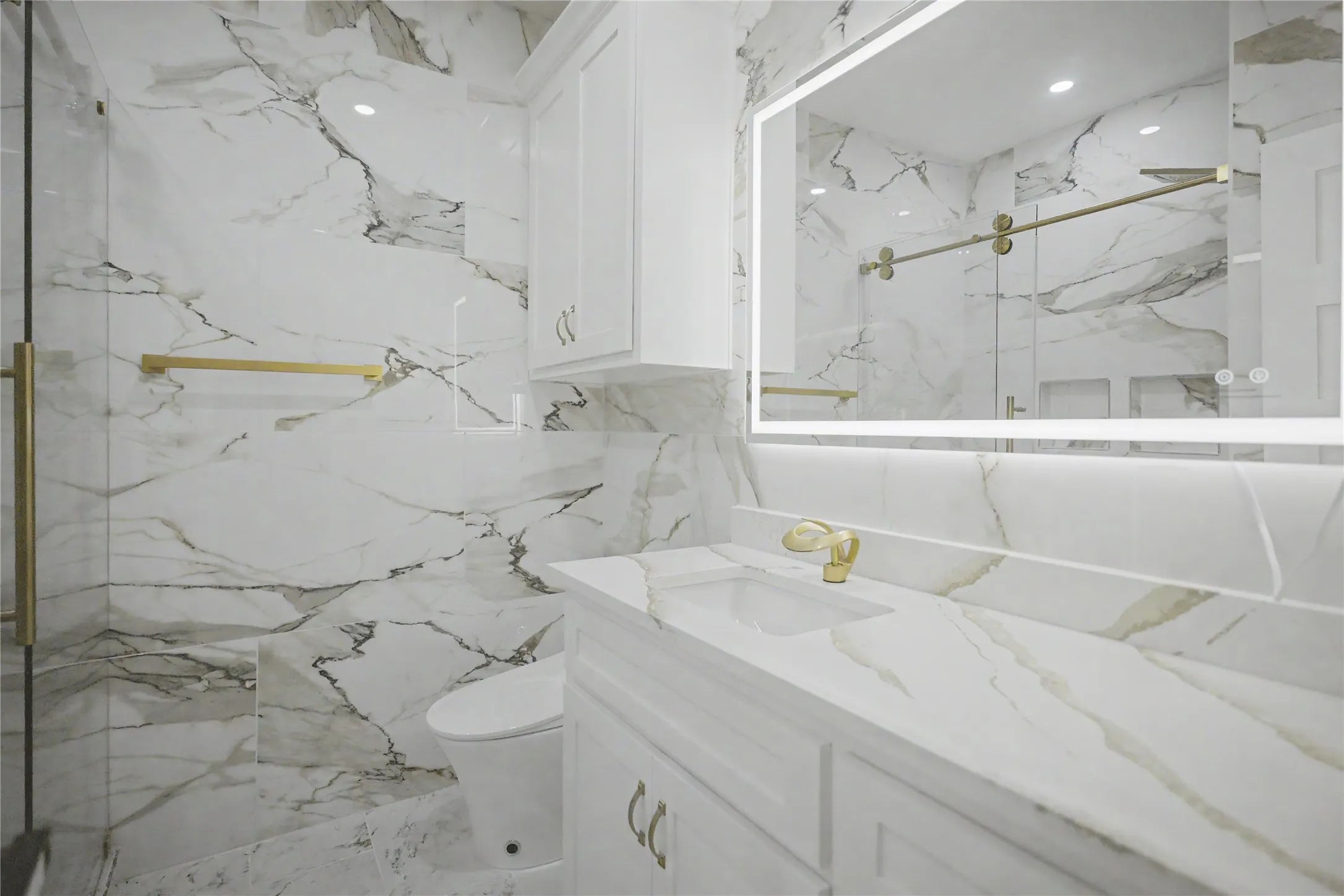 Bathroom featuring floor-to-ceiling marbled wall and floor tiles, a glass-enclosed shower with gold-tone hardware, a white vanity with a marbled countertop, and an illuminated mirrored cabinet
