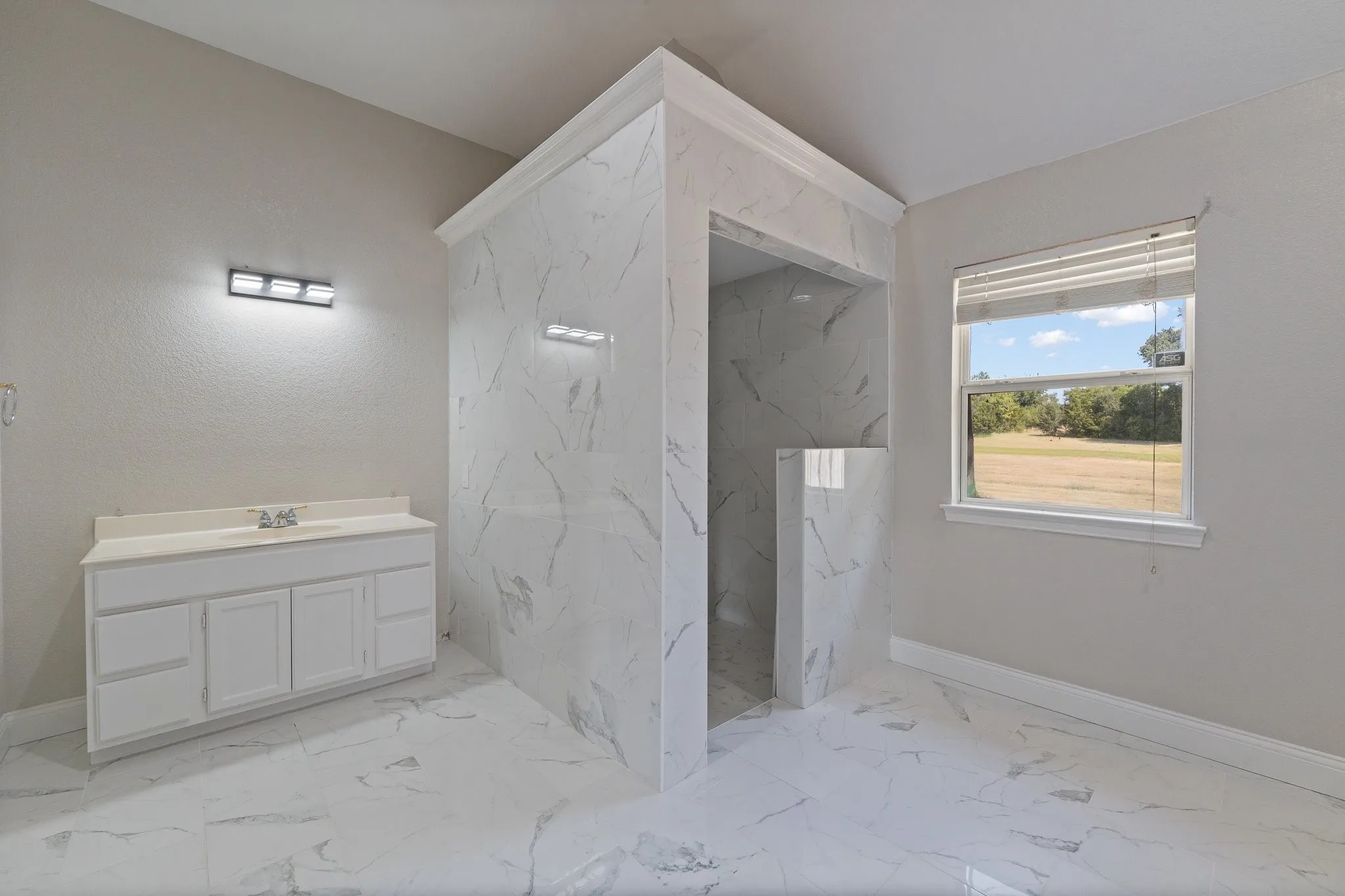 Walk-in shower with white marbled tile, white vanity with integrated sink, contemporary wall-mounted light fixture, and white window blinds