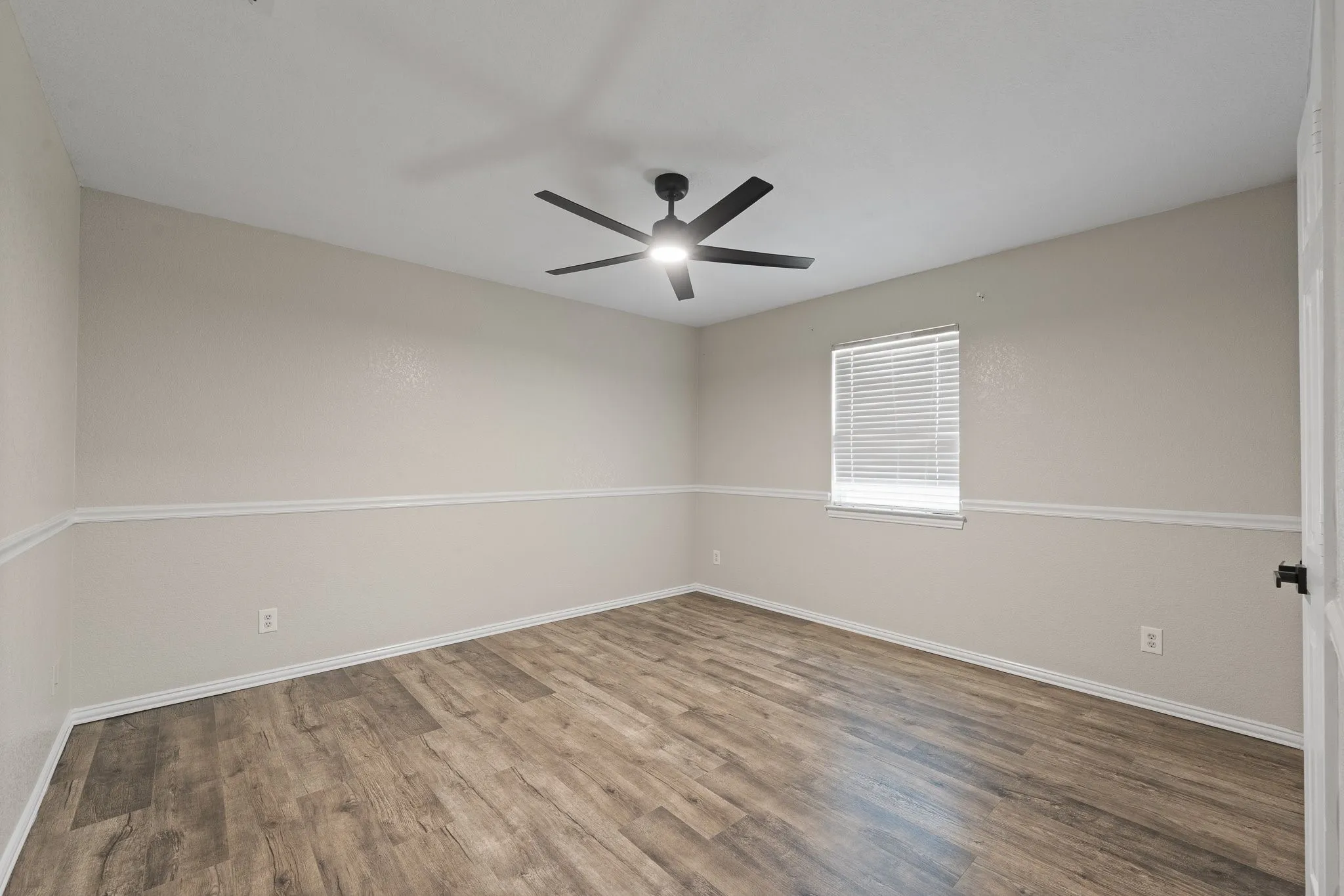 Spacious room featuring wood-finish flooring, two-tone wall paint with decorative trim, a window with blinds, and a contemporary ceiling fan with integrated lighting