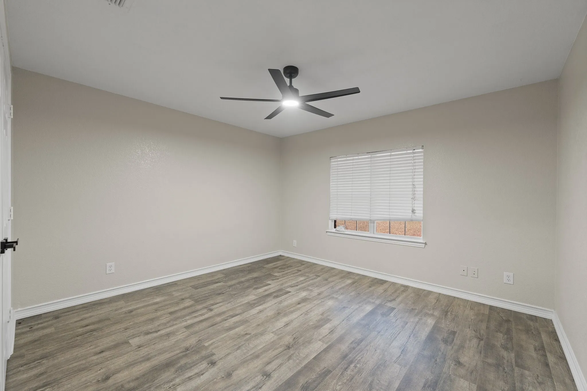 Spacious room featuring wood-finish flooring, light-toned walls, white trim, a modern black ceiling fan with integrated lighting, and a window with blinds