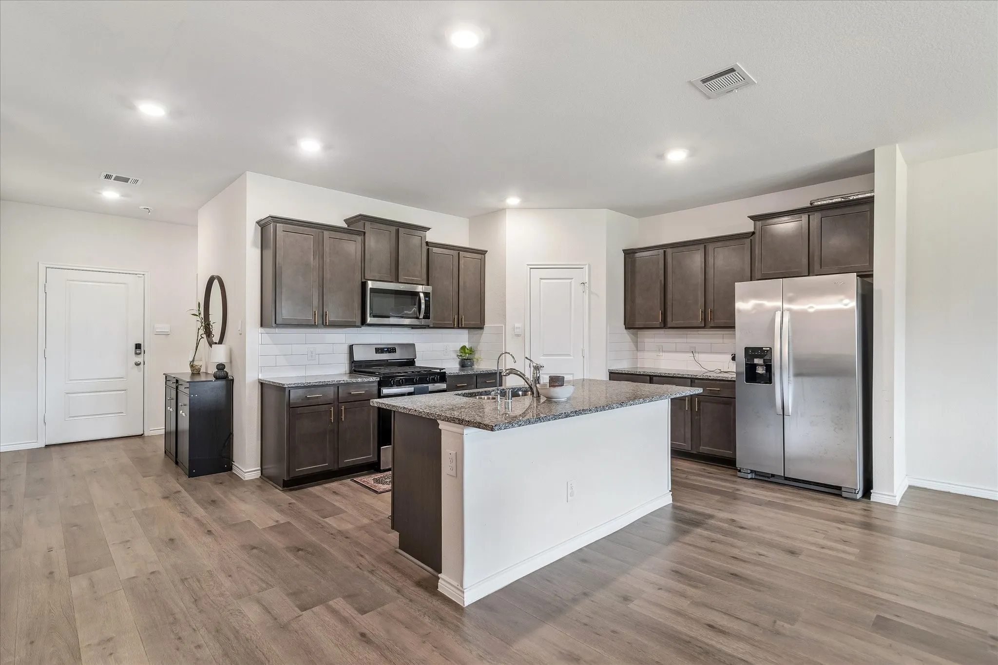 Open-concept kitchen featuring an island with a double basin sink, dark wood-finish cabinetry, stainless steel appliances, subway tile backsplash, and wood-finish flooring