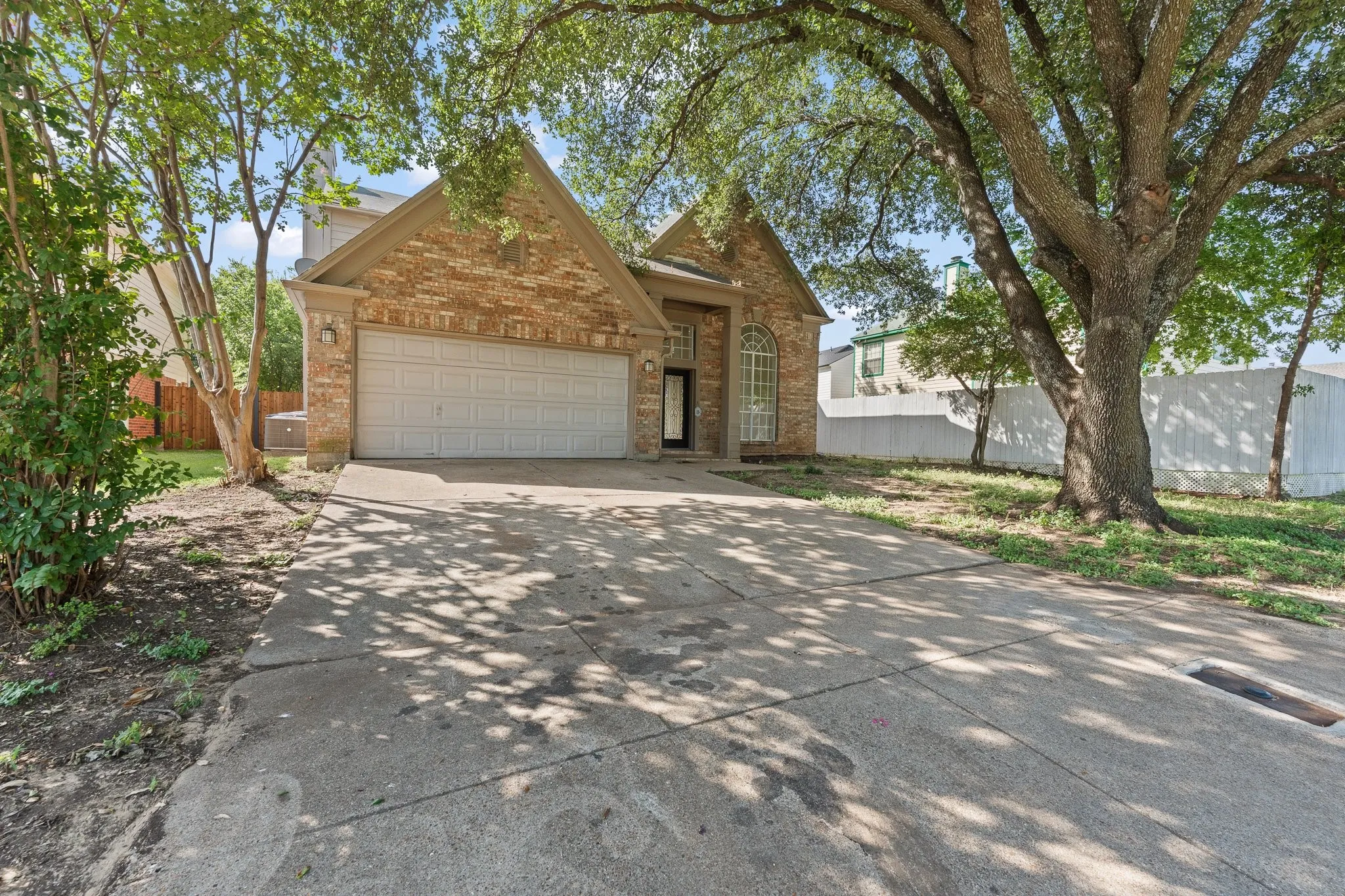Brick facade residence featuring a front-facing garage, arched entry window, and mature front yard trees