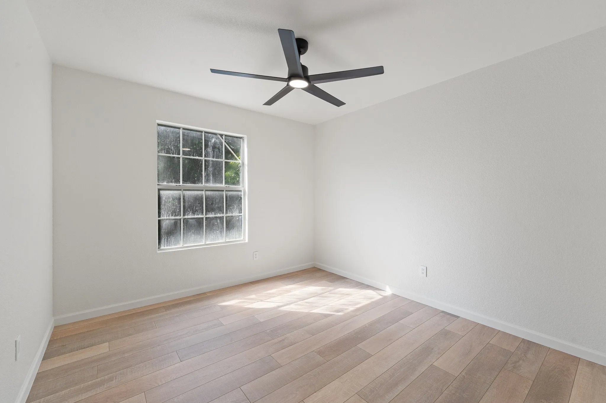 Spacious room featuring light wood-finish flooring, a modern black ceiling fan with integrated lighting, a large window with multiple panes, and crisp white walls