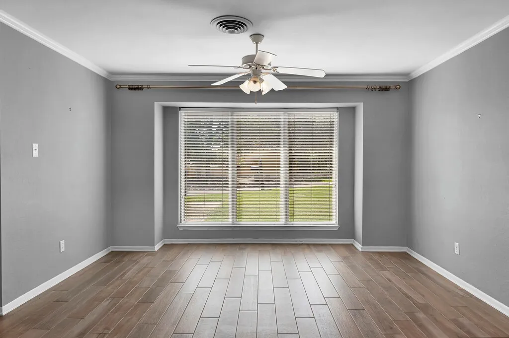 Spacious room featuring wood-finish tile flooring, a large window with blinds, a ceiling fan with light fixture, crown molding, and baseboards