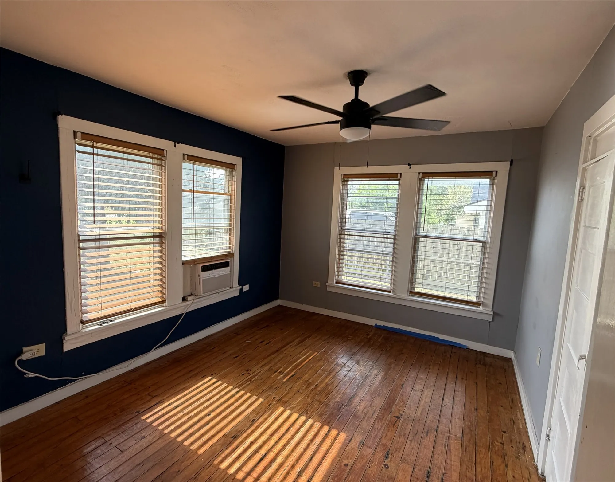 Room featuring wood-finish flooring, a ceiling fan with integrated lighting, multiple windows with blinds, and a window-mounted air conditioning unit