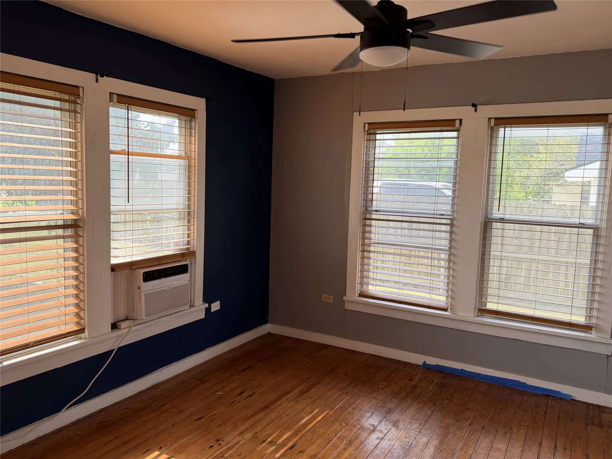 Room featuring three windows with wood-finish blinds, wood-finish flooring, a ceiling fan with integrated lighting, and walls with contrasting blue and gray paint