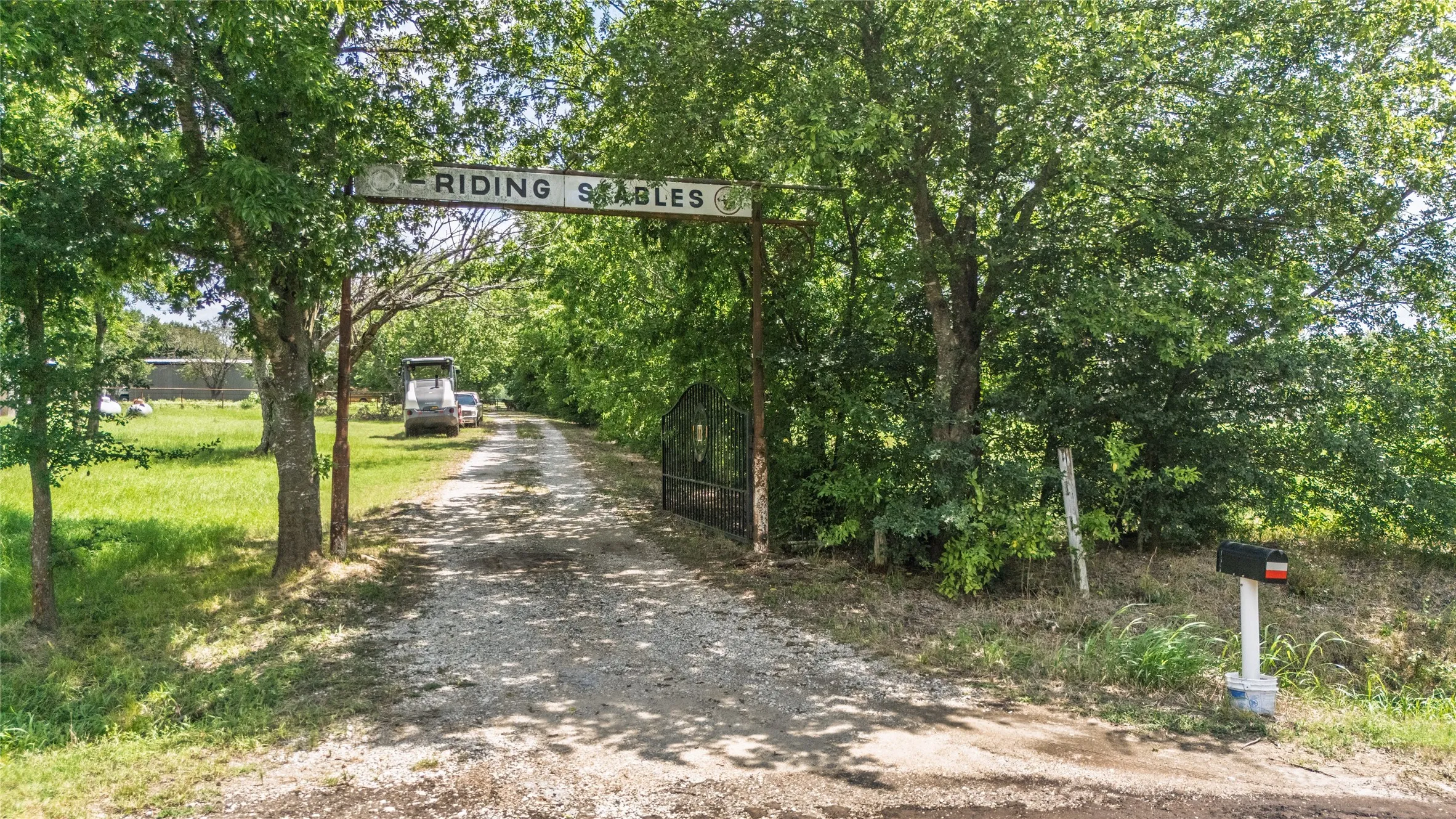 Gated entrance featuring a "Riding Stables" sign, established trees, and a gravel driveway