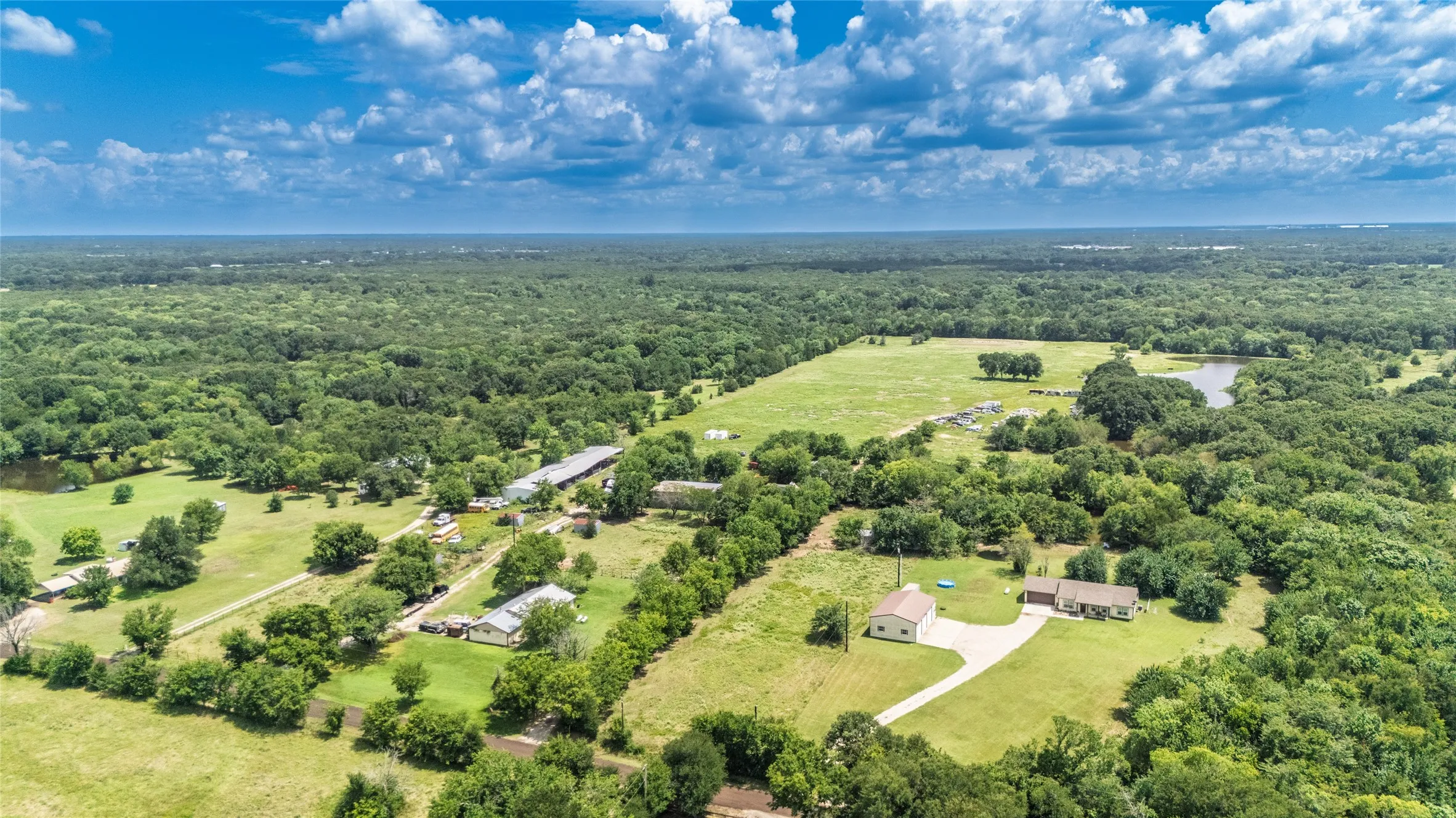 Expansive aerial landscape featuring multiple structures, verdant fields, wooded areas, and a natural pond