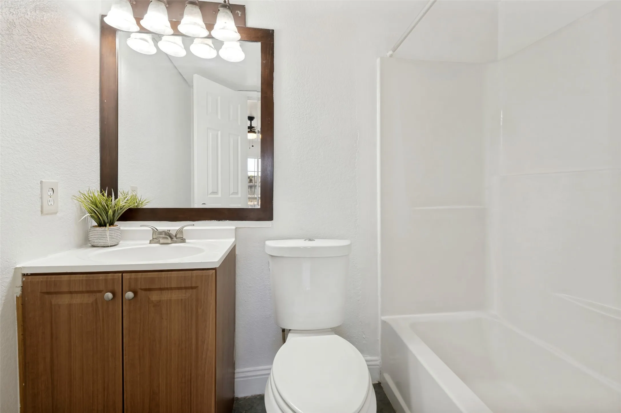 Bathroom featuring a wood-finish vanity with an integrated sink and chrome faucet, a framed mirror with a four-bulb light fixture, a toilet, and a white bathtub with a shower surround