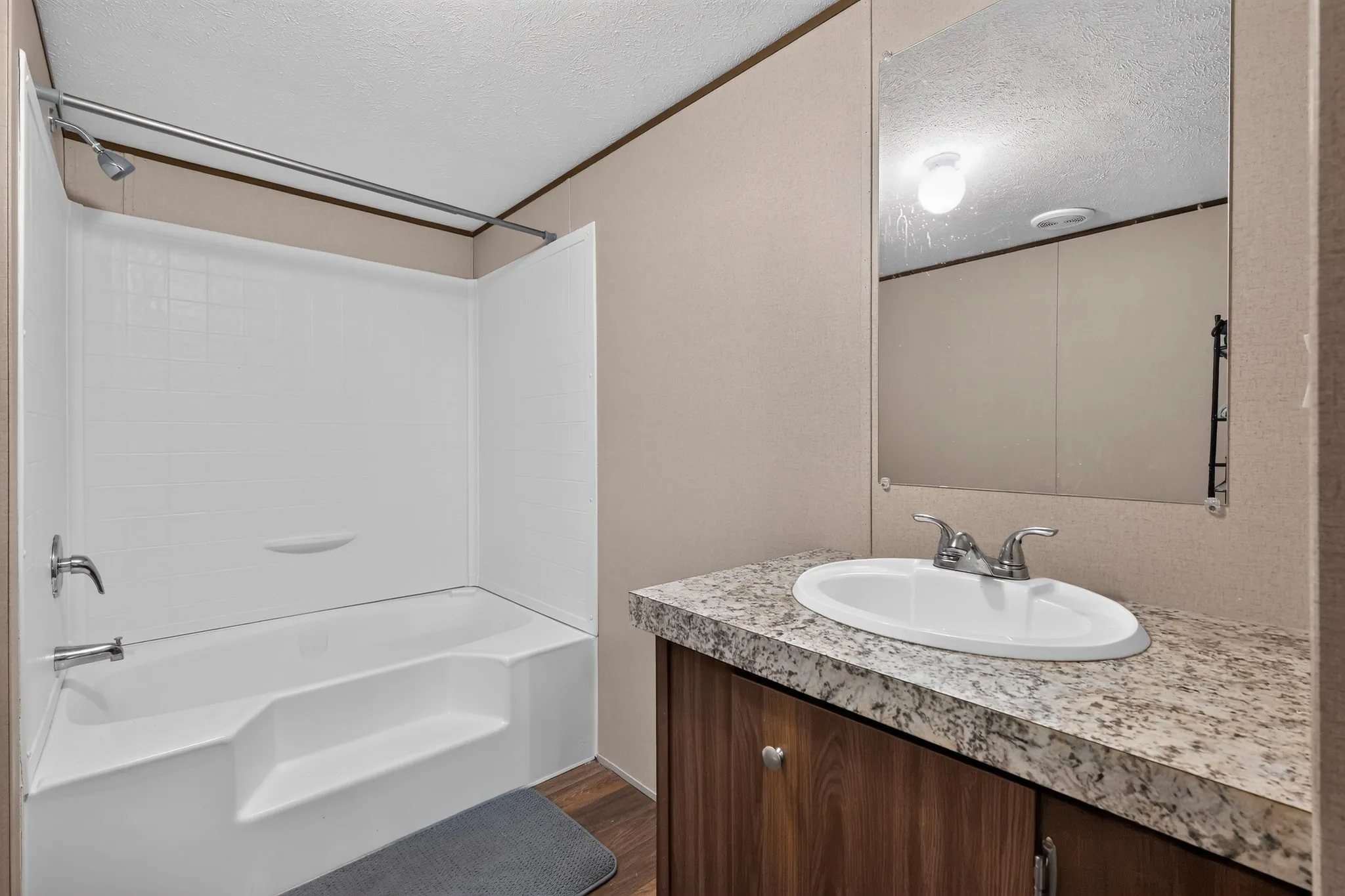 Full bathroom featuring a white tub/shower combination, chrome fixtures, a wood-finish vanity with a speckled countertop, and a wall-mounted mirror