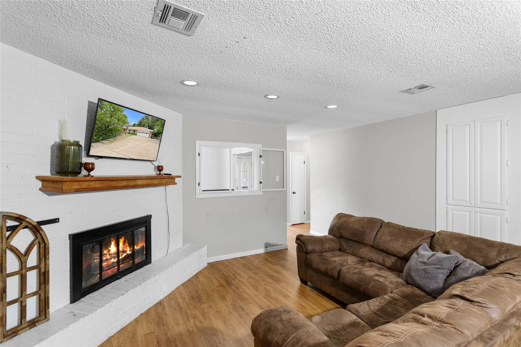 Living area featuring a painted brick fireplace with a wood mantel, wood-finish flooring, recessed lighting, and built-in cabinetry