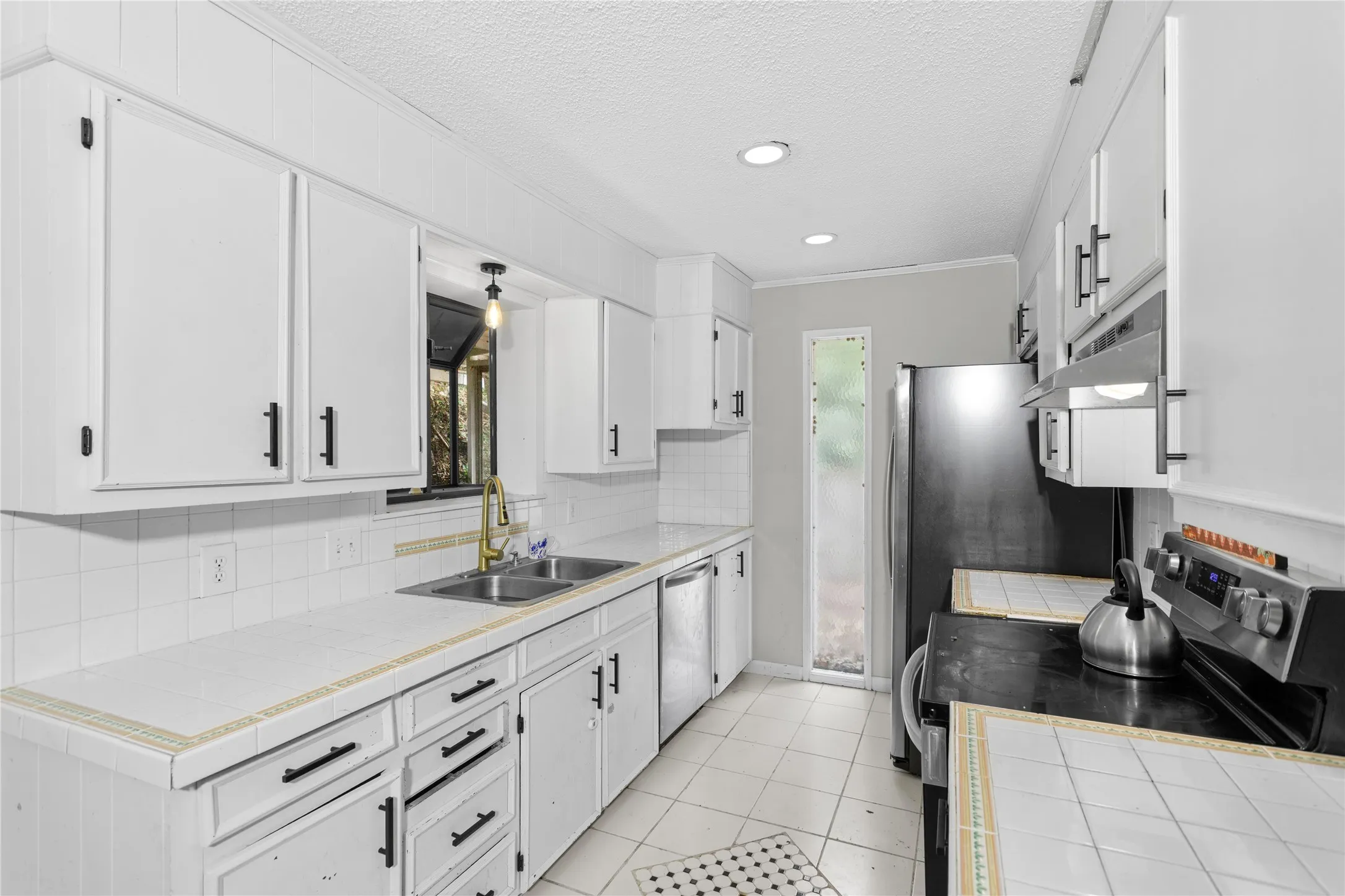 Kitchen featuring white cabinetry with black hardware, tile countertops, stainless steel double basin sink with a gold-tone faucet, and white tile flooring