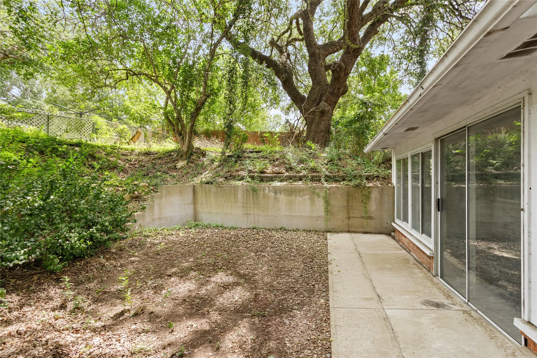 Rear exterior patio featuring concrete pavers, a substantial retaining wall, and a large mature tree