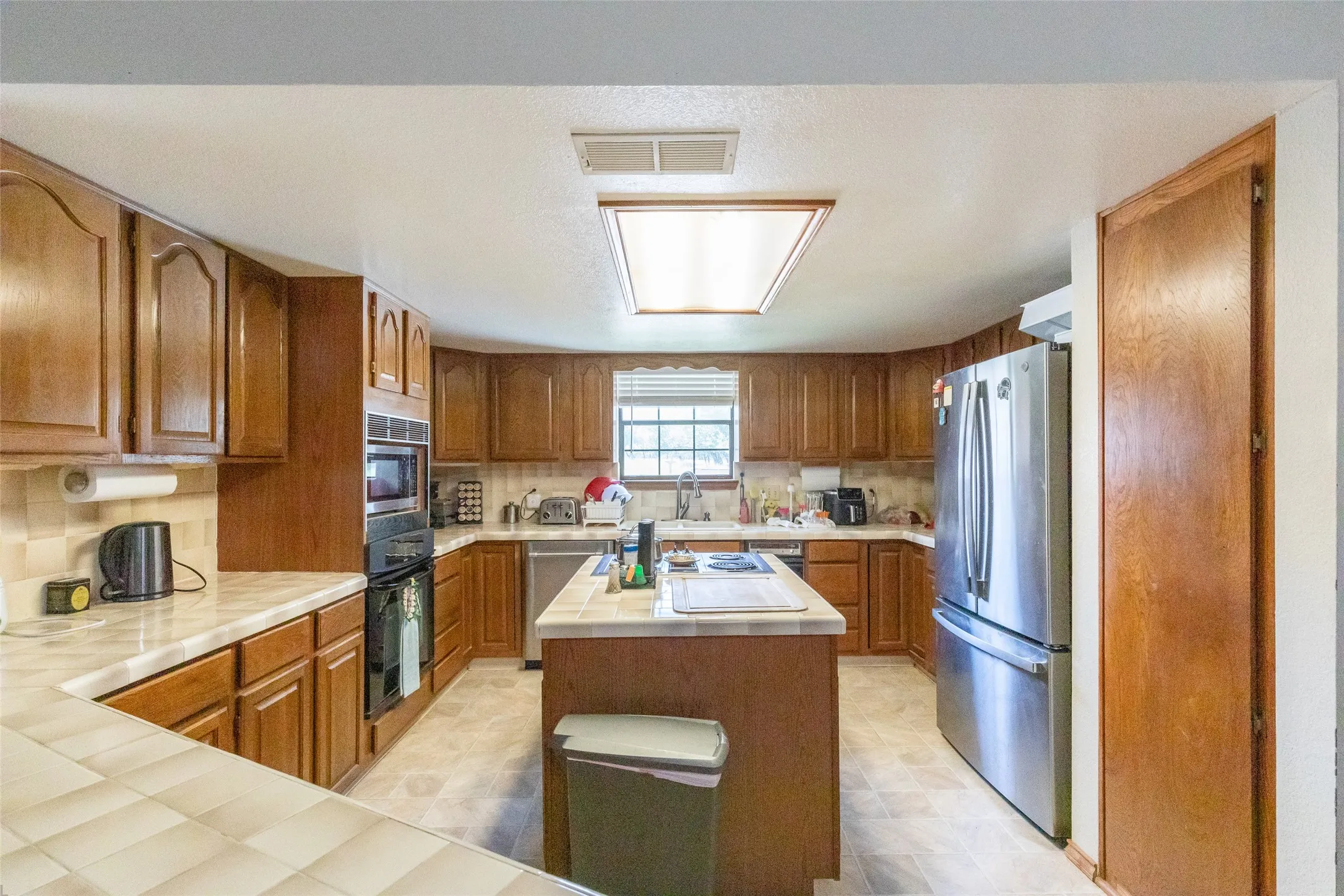 Kitchen featuring wood cabinetry, tiled countertops, a central island, built-in double oven, and stainless steel refrigerator