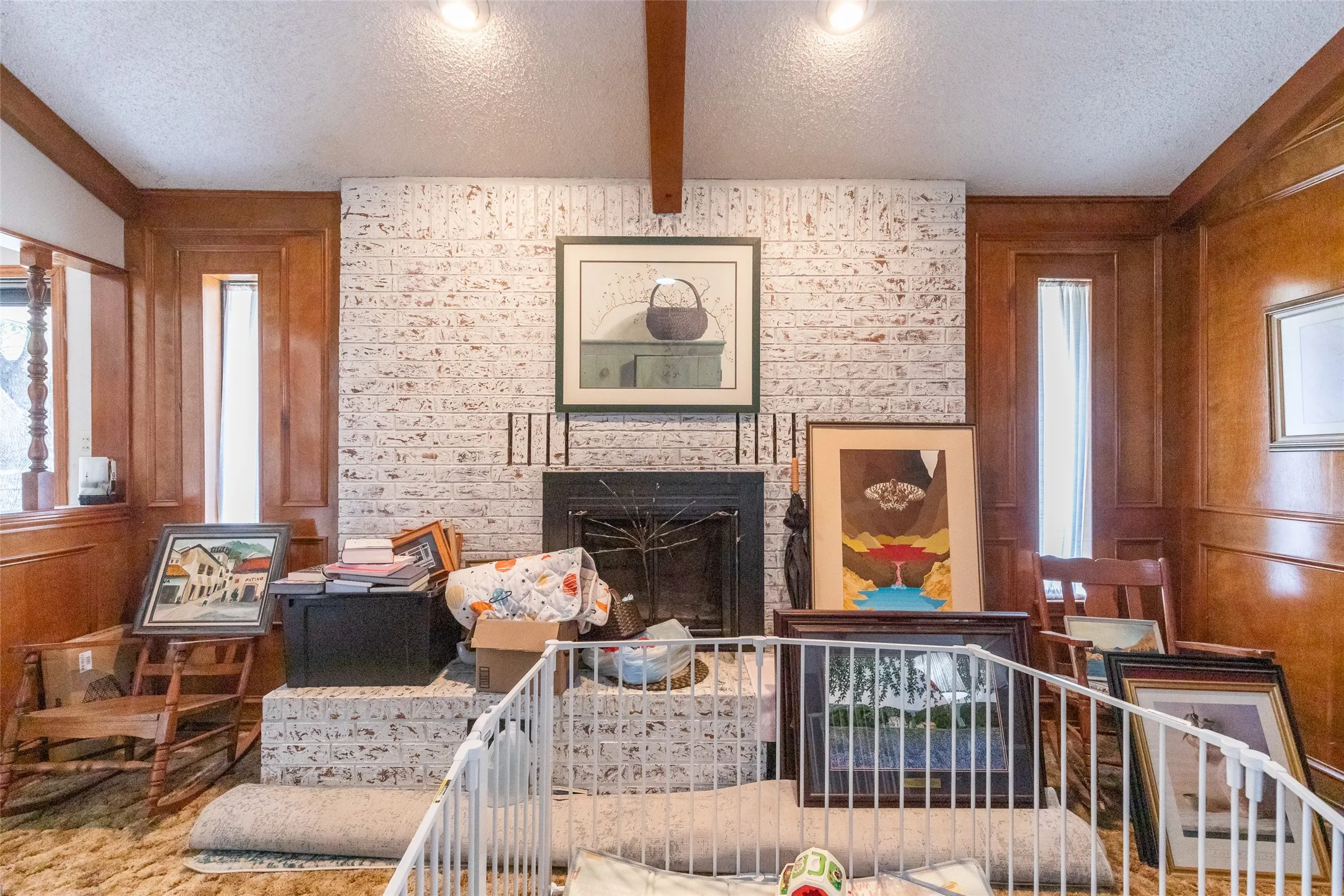 Whitewashed brick fireplace with a dark stone hearth, wood-paneled walls, exposed ceiling beam, and recessed lighting