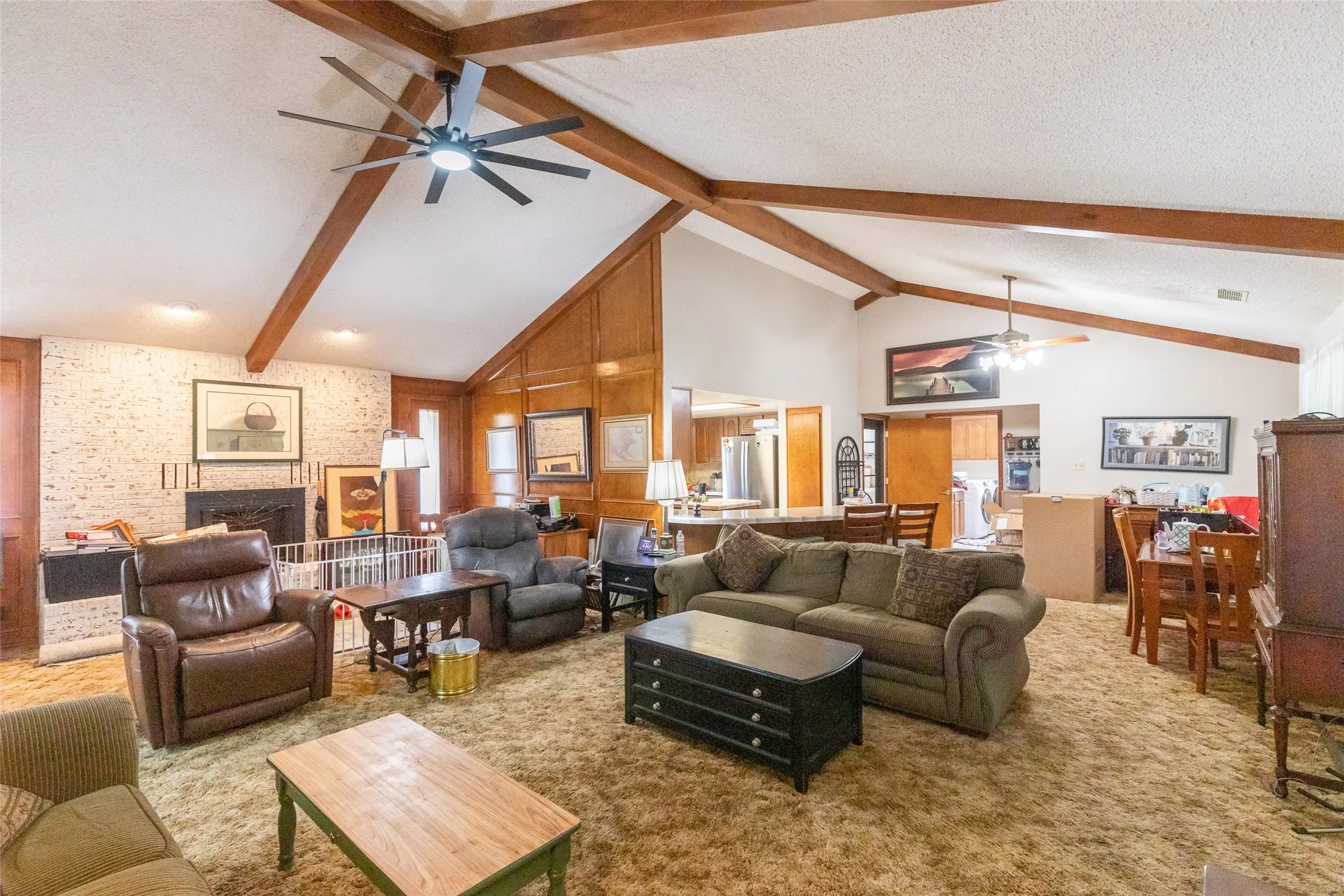Spacious living area featuring a vaulted ceiling with exposed wood beams, a white brick fireplace, wood-paneled walls, and a ceiling fan with integrated lighting