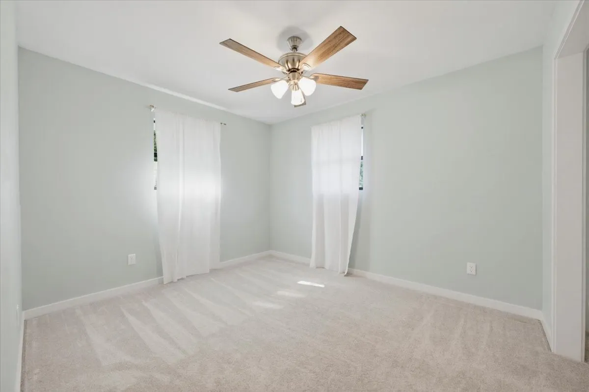 Light-colored carpeted flooring, neutral wall tones, two windows with white sheer curtains, a ceiling fan with wood-finish blades, and white baseboards