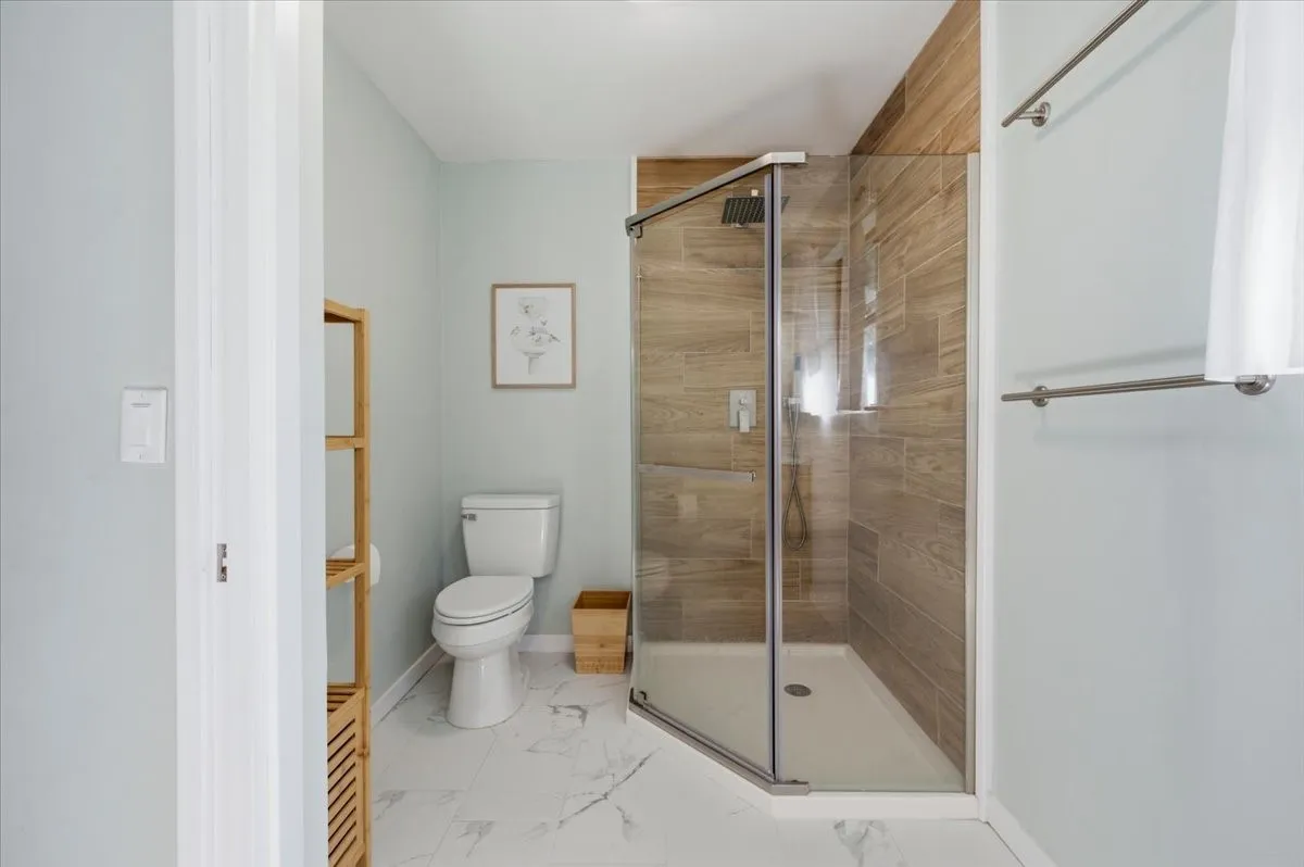 Modern bathroom featuring a glass-enclosed shower with wood-finish tile surround, white porcelain toilet, and marble-look floor tiling