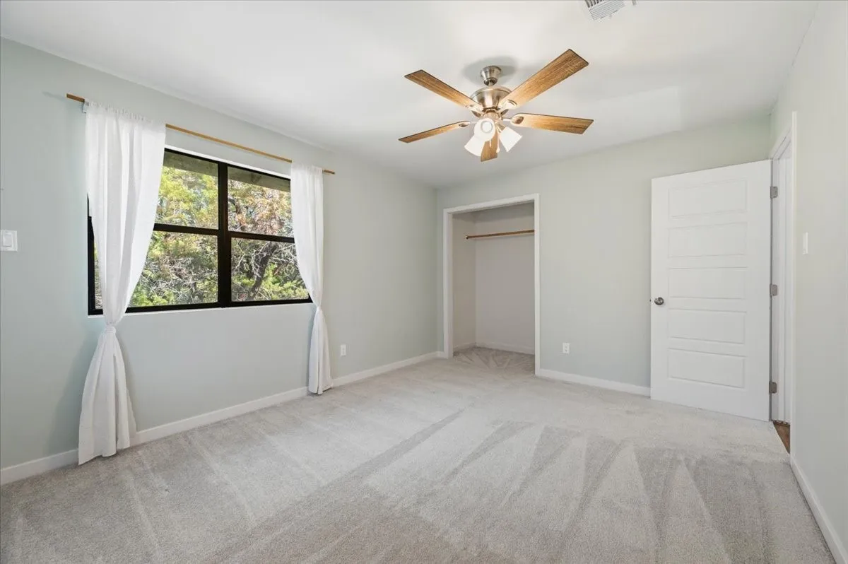Spacious room featuring light-toned carpeting, a ceiling fan with wood-finish blades, and a large window with white curtains