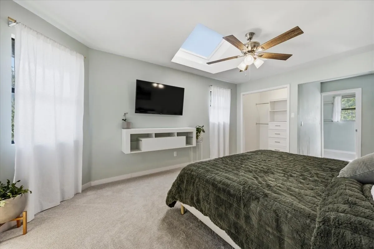 Vaulted ceiling with skylight and ceiling fan, built-in closet system with drawers and shelving, wall-mounted media console, and light-colored carpeting