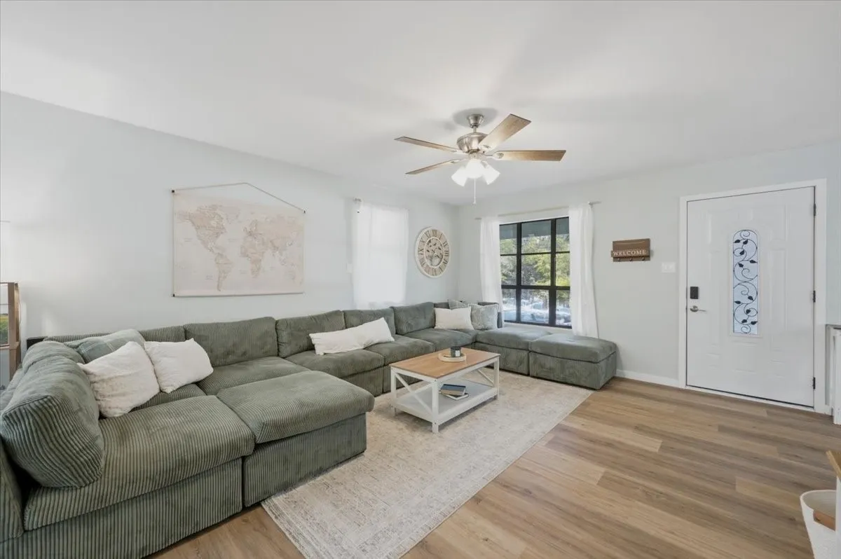 Living area featuring light-toned walls, wood-finish flooring, a ceiling fan, a window with dark trim, and a white entry door with decorative glass